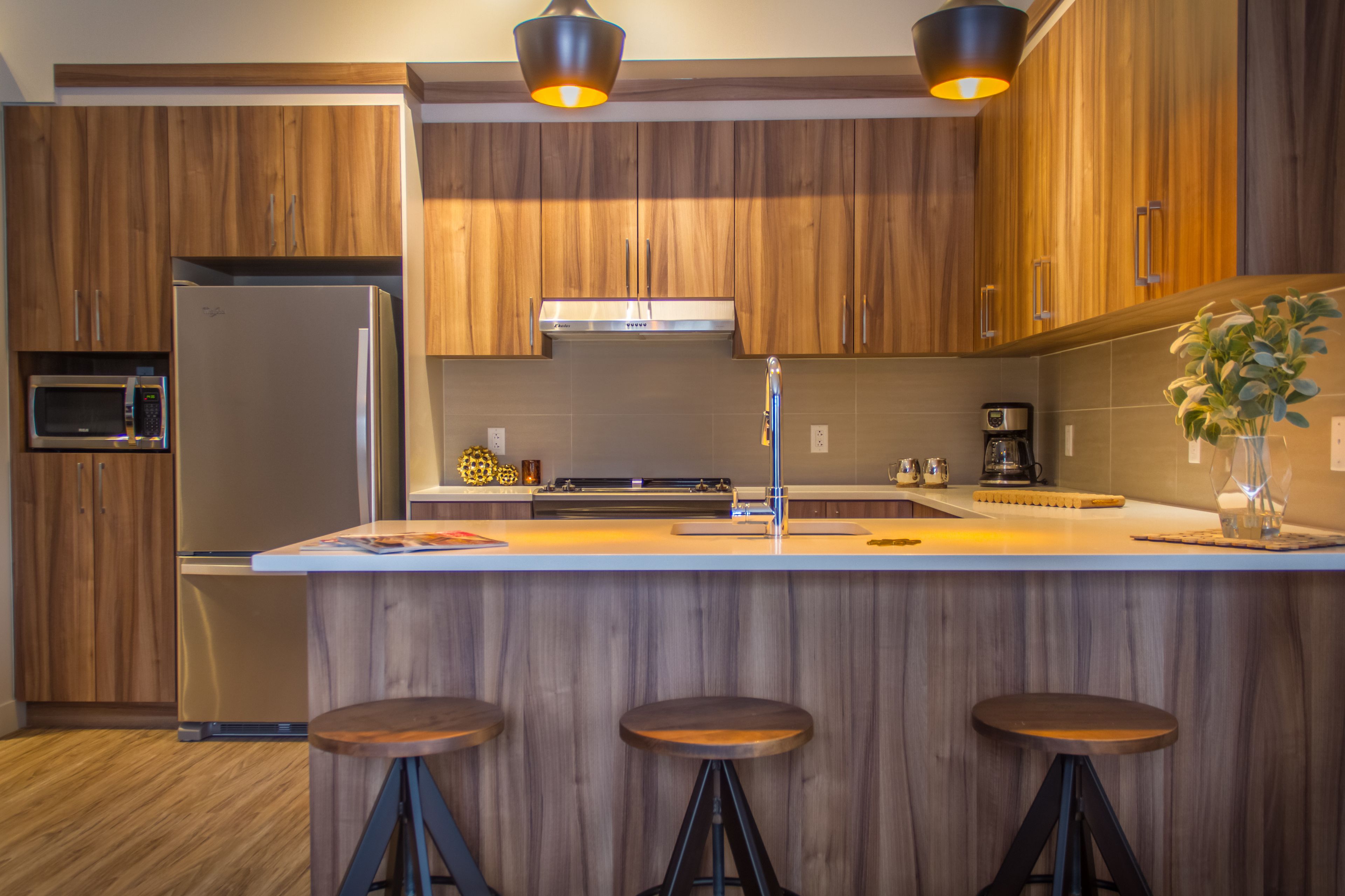 Une cuisine moderne avec des armoires en bois, un réfrigérateur en acier inoxydable, un four à micro-ondes et une machine à café. Il y a trois tabourets au niveau de l’îlot central.
