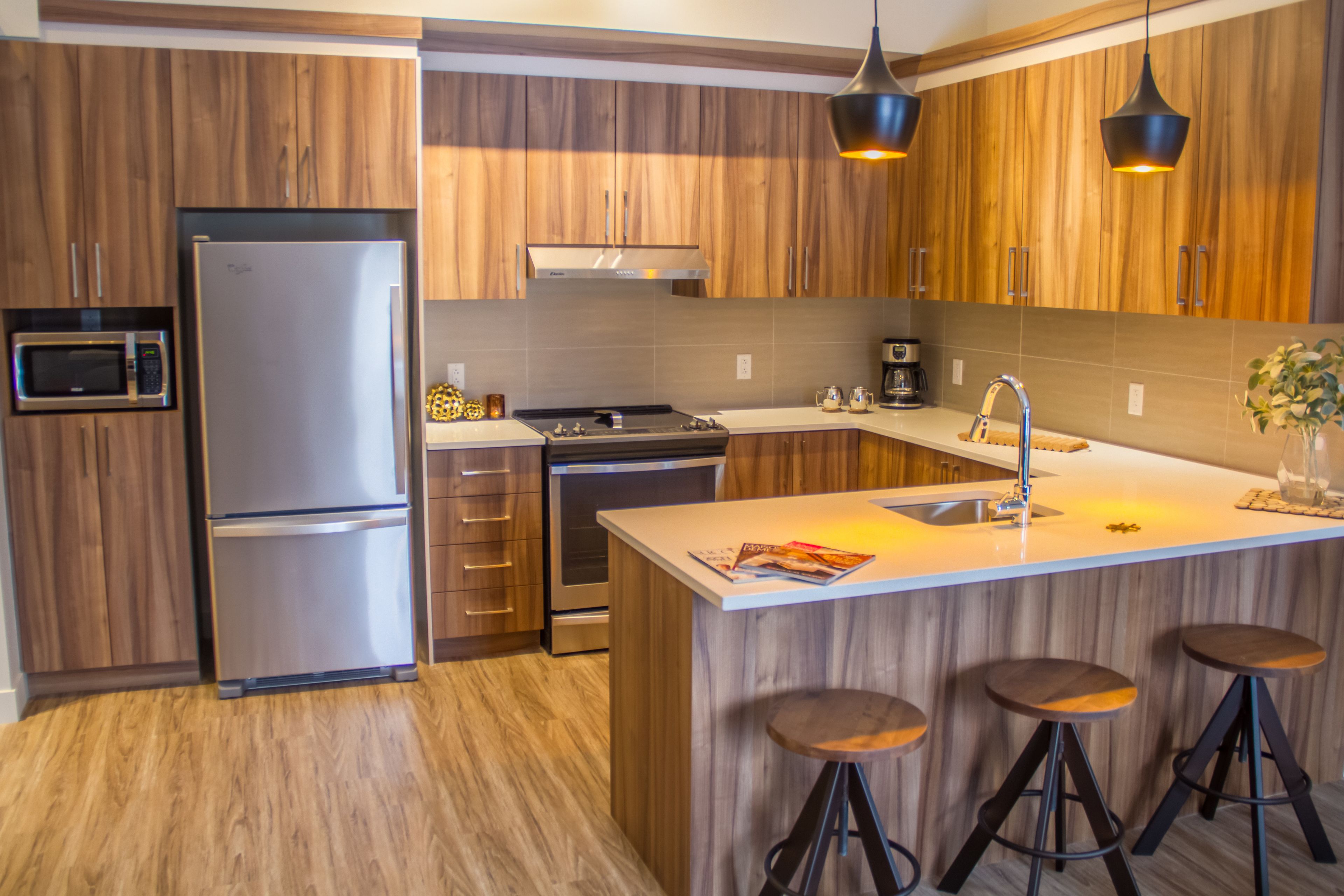 A modern kitchen with wooden cabinetry, stainless steel appliances, a central island with stools, and pendant lighting.