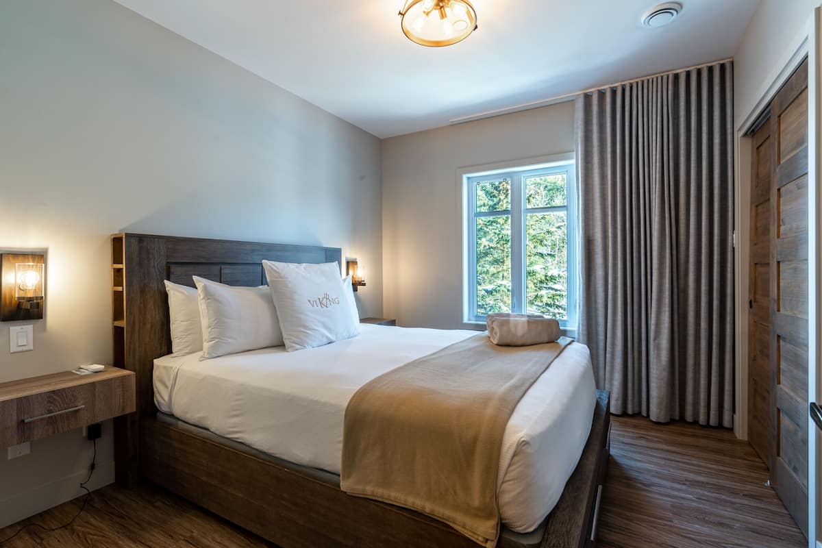Un lit bien fait, avec une tête de lit, une table de chevet avec une lampe et une fenêtre avec des rideaux.