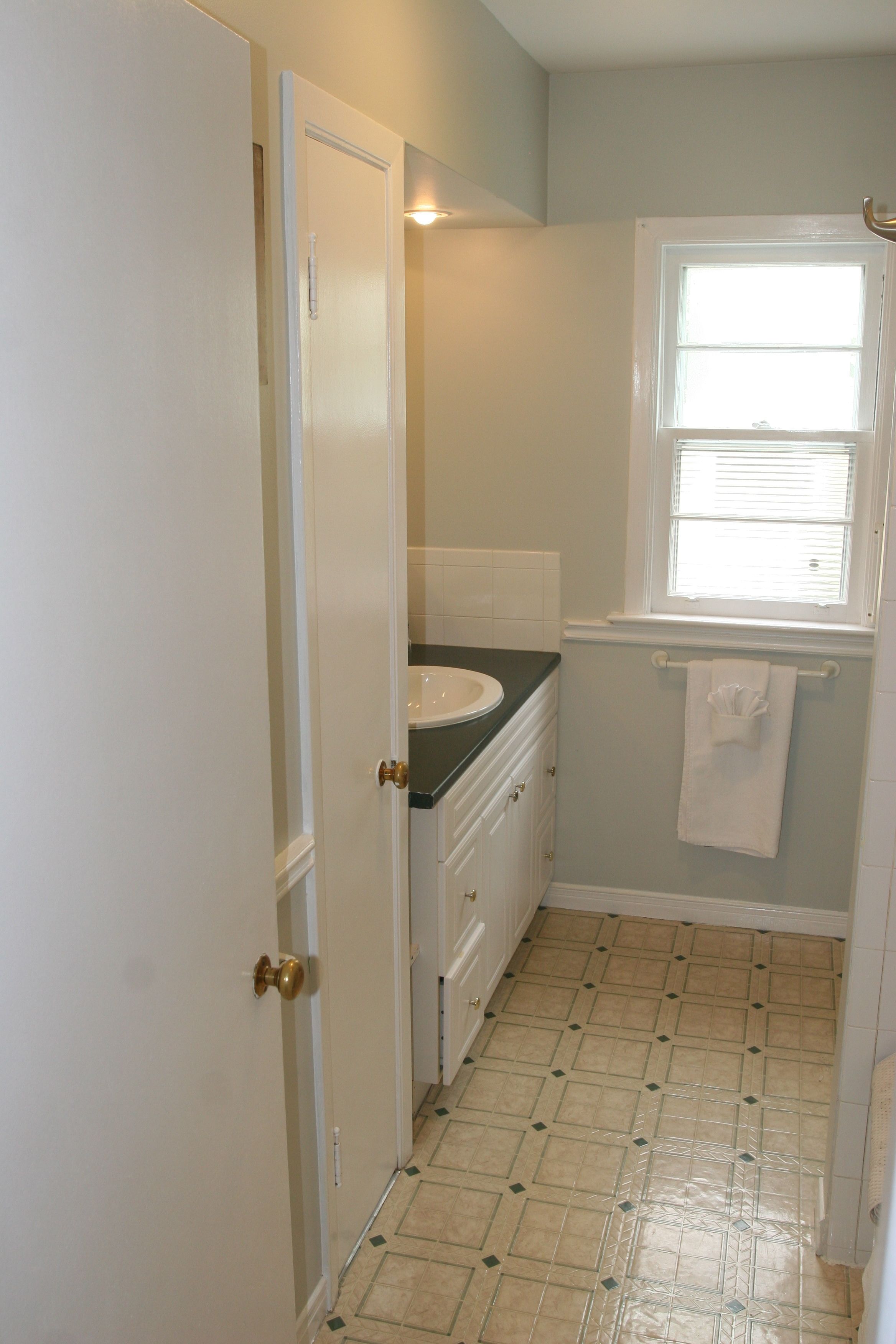 A bathroom with a white door, a sink, a window with blinds, and a tiled floor.