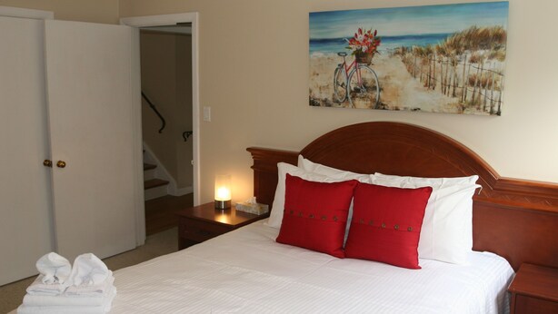A neatly made bed with white and red pillows, a painting of a beach scene on the wall, and a wooden headboard. - Niagara Riverview Inn (Niagara Falls)