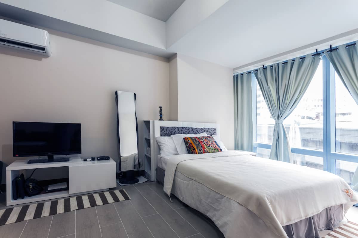 A modern hotel room with a flat-screen TV, a bed with a white headboard, a large window with curtains, and a small desk with a mirror.