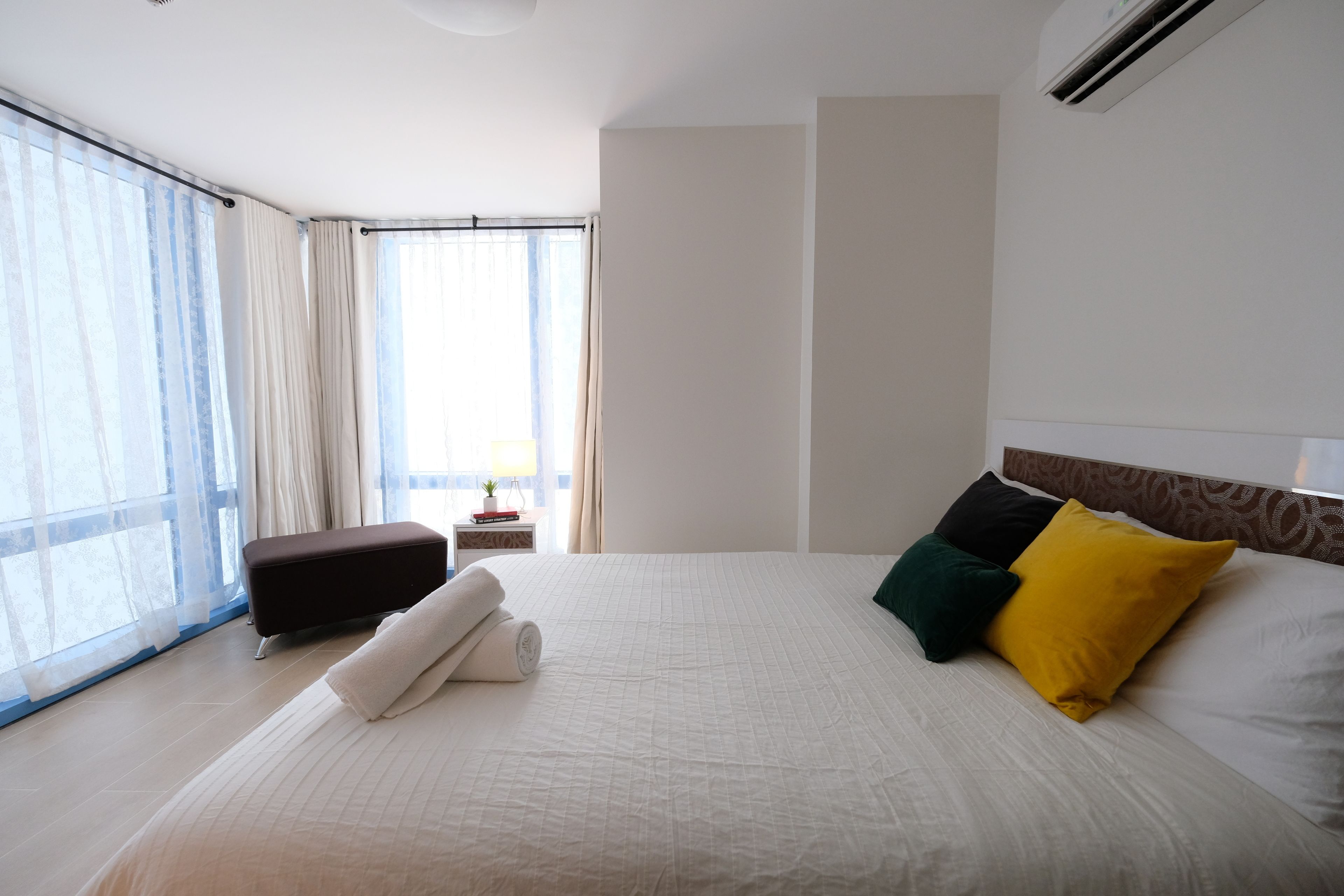 A hotel room with a large bed, a small bedside table, a window with sheer curtains, and a view of the outdoors.