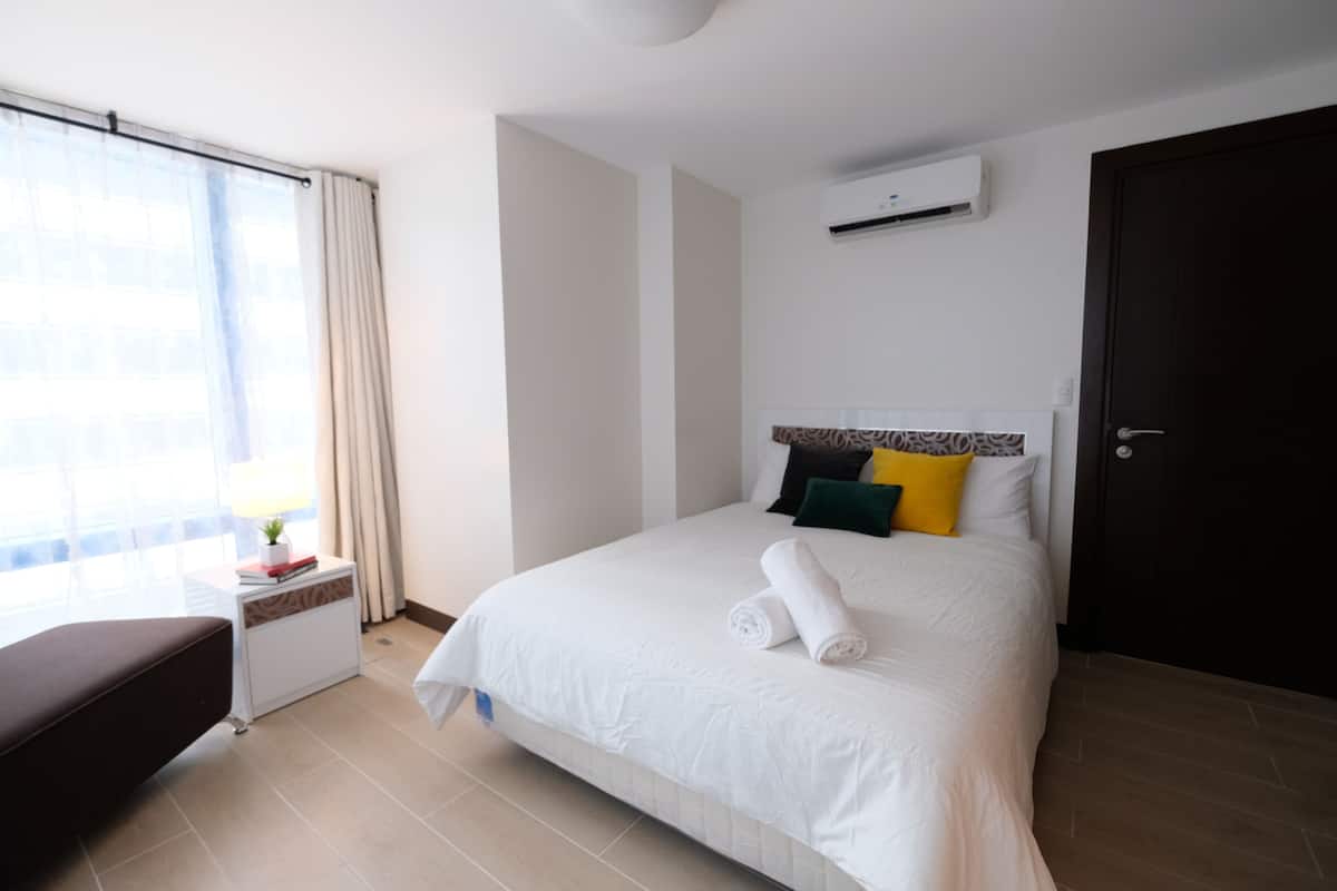 A hotel room with a bed, a bedside table, a window with curtains, an air conditioning unit, and a door.