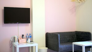 Una habitación con un televisor de pantalla plana negro montado en la pared, una mesa blanca con una tetera, una taza y una caja con el rótulo "kiss you", un área de descanso con cojines negros y una mesita blanca con una revista.