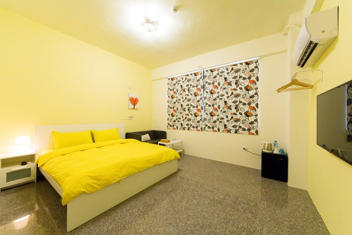 Un dormitorio con cama, mesita de noche, cómoda, televisor de pared y ventana con estampado floral.