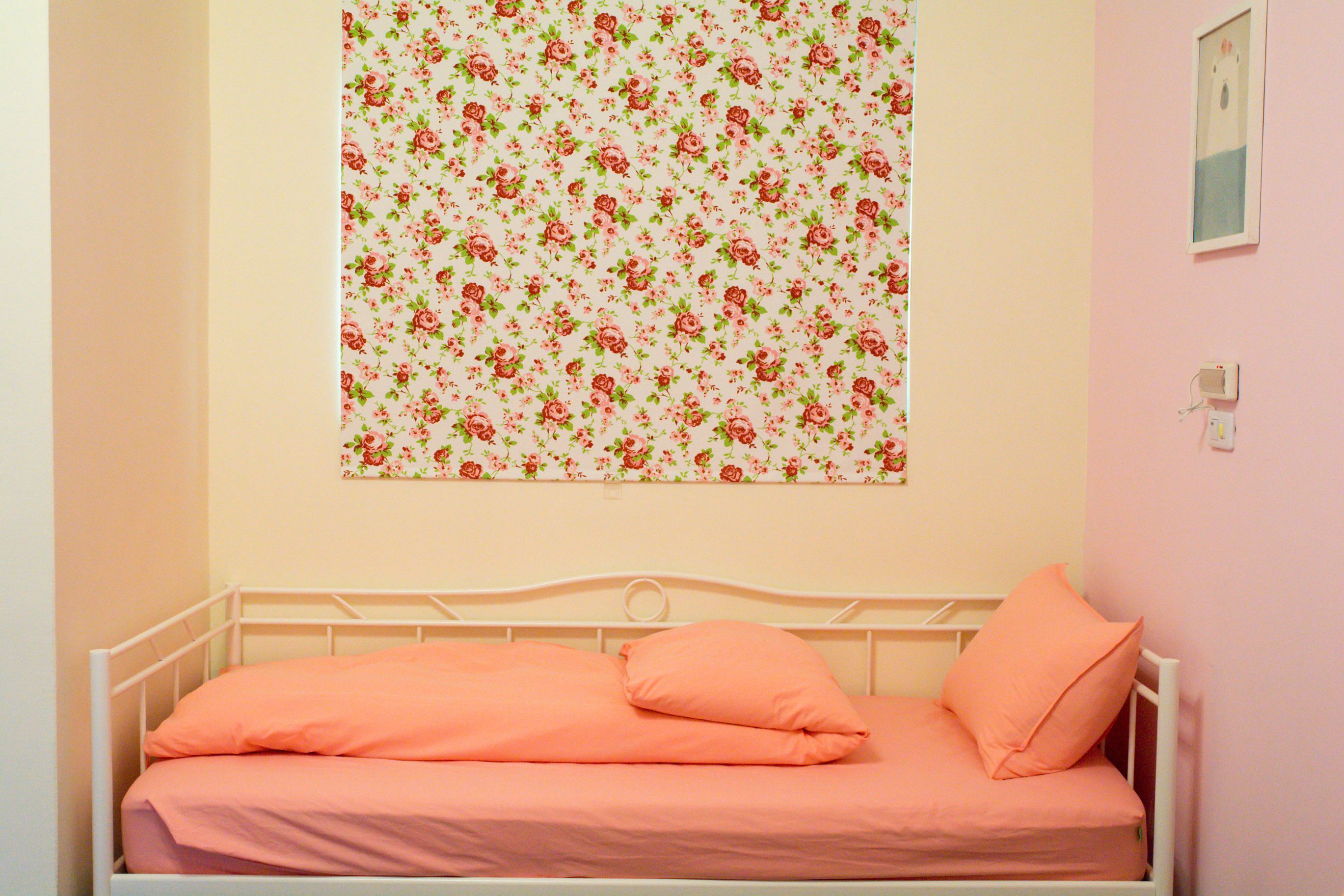 Una cama individual con estructura metálica blanca y ropa de cama color melocotón en una habitación con paredes rosa claro.