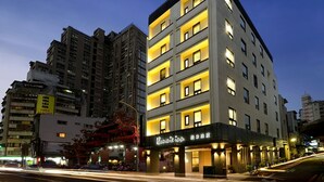 A multi-story hotel with illuminated balconies, a traditional red building with a dragon decoration, and a street with moving vehicles. - Hommie Inn (Taichung)