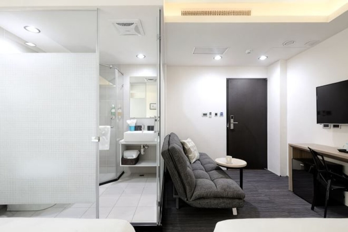 A modern hotel room with a sofa, a desk, a TV, a bathroom with a shower, and a door.