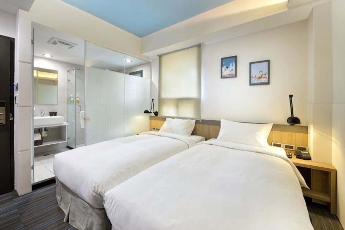 A hotel room with two beds, a wardrobe, a mirror, a bedside table with a lamp, and a small shelf.