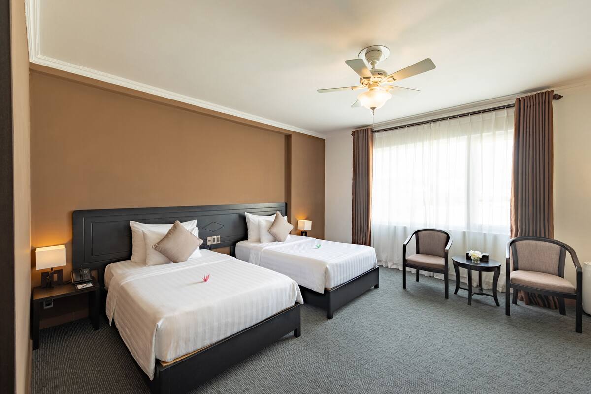 Une chambre d’hôtel avec deux lits, un ventilateur de plafond, une petite table avec un vase et deux chaises.