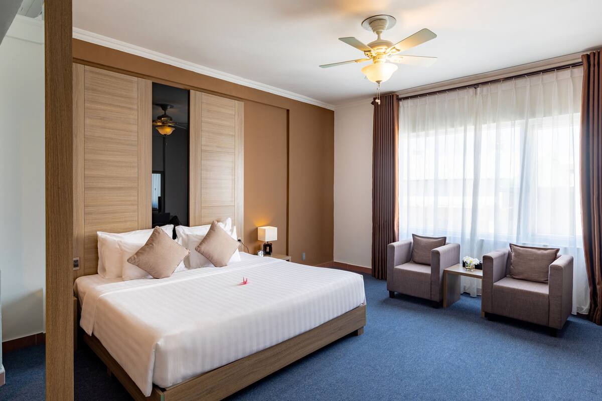 Une chambre d’hôtel avec un grand lit, deux fauteuils, une petite table, un ventilateur de plafond et une grande fenêtre avec des rideaux.