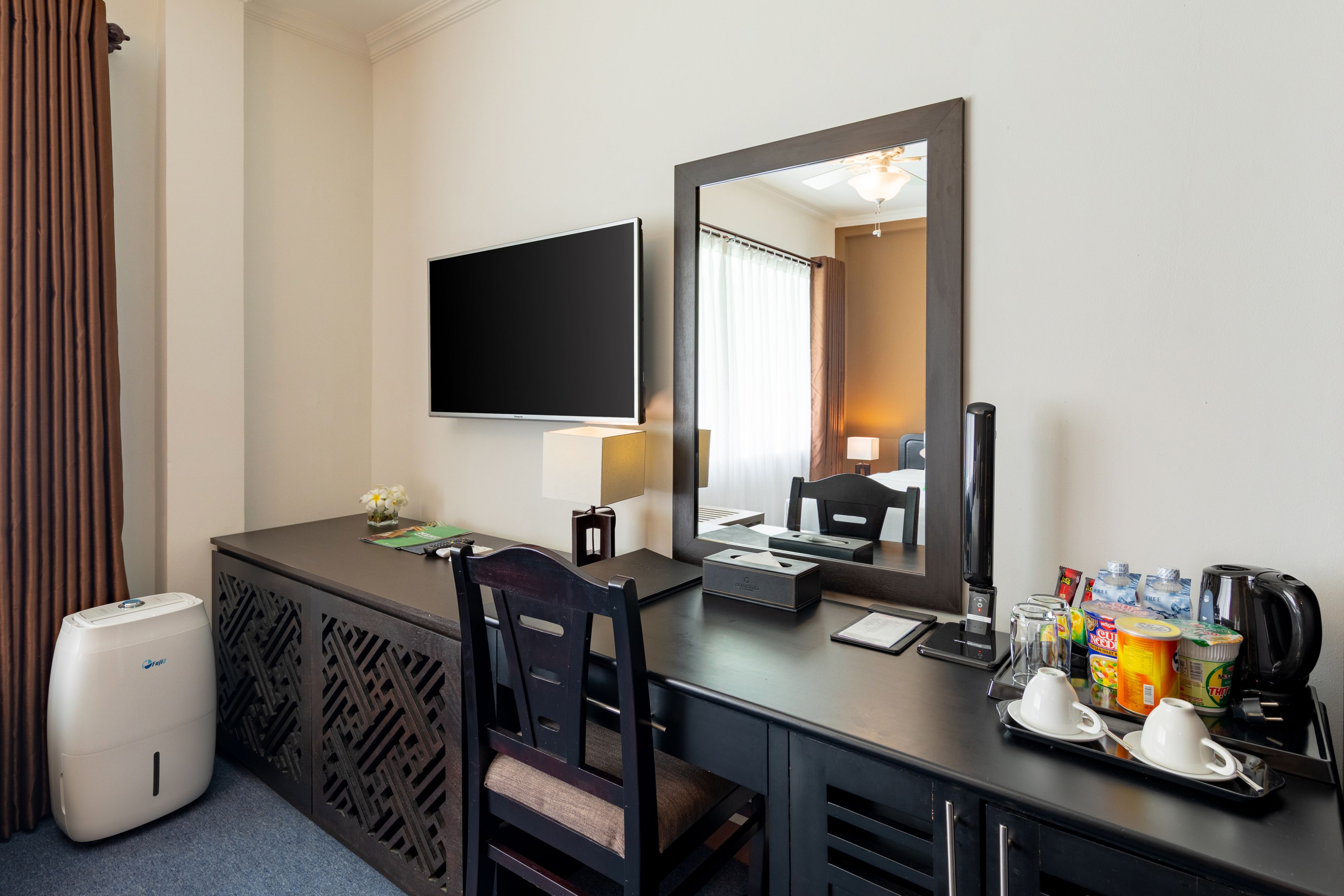 A hotel room with a desk, a flat-screen TV, a mirror, a coffee maker, and a trash can.