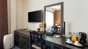 A hotel room with a desk, a flat-screen TV, a mirror, a coffee maker, and a trash can.