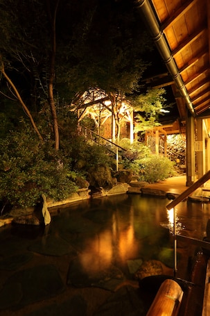 A traditional Japanese garden at night with a wooden structure, a pond, and surrounding vegetation.