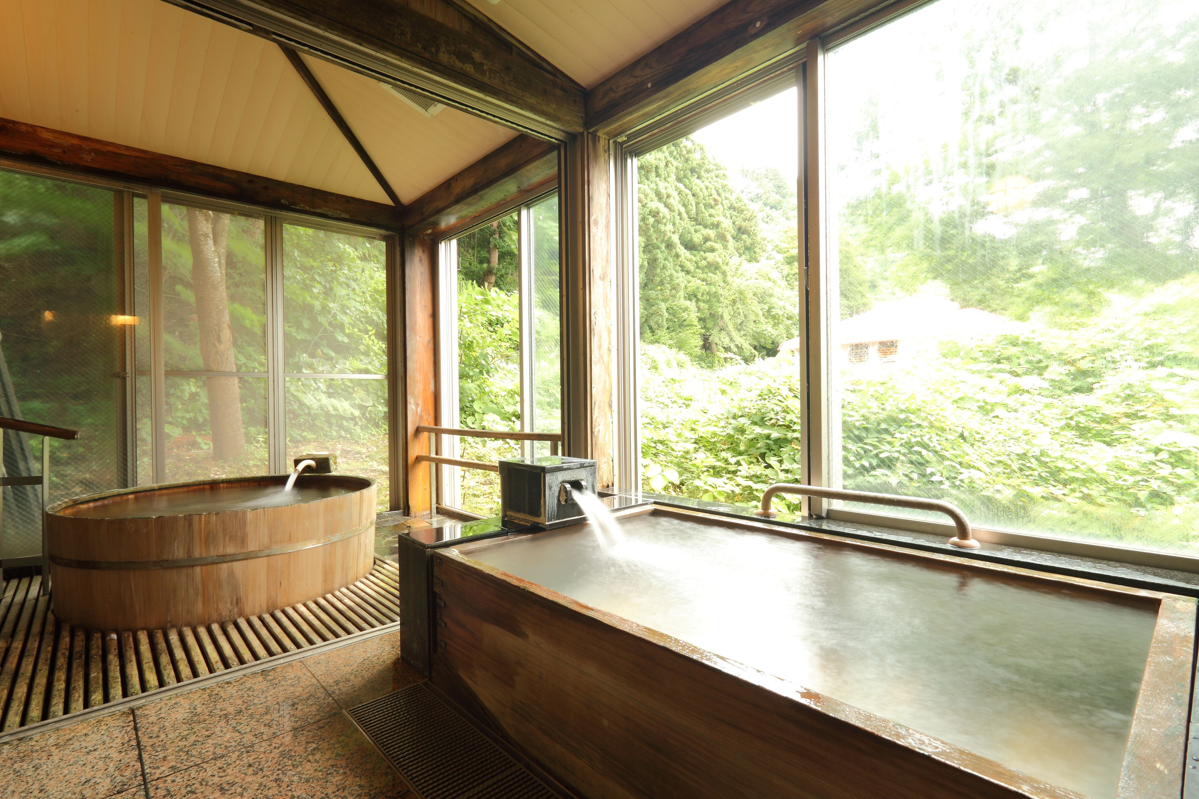 A wooden bathtub with a faucet, large windows, and a view of a forested area.