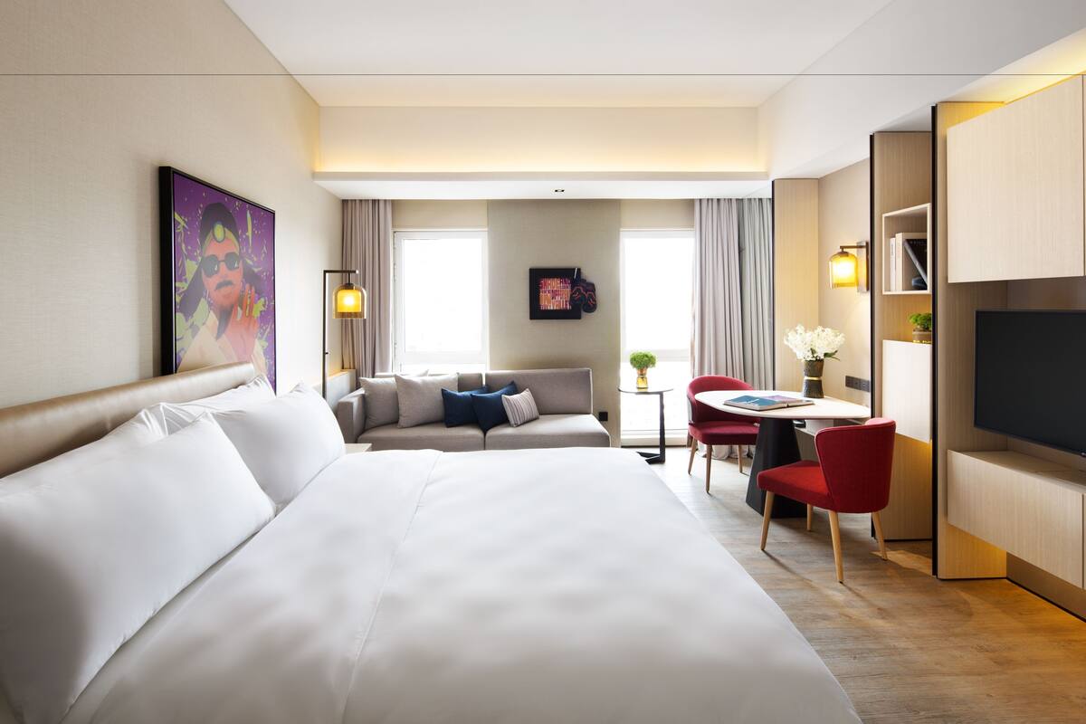 A hotel room with a large bed, a sofa, a desk with a chair, a television, and a painting on the wall.