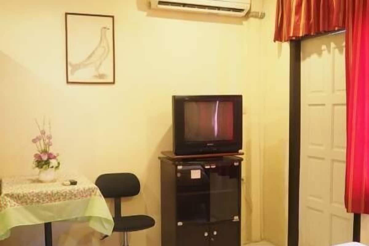 A hotel room with a bed, a desk with a chair, a television on a cabinet, an air conditioner, and a door with red curtains.