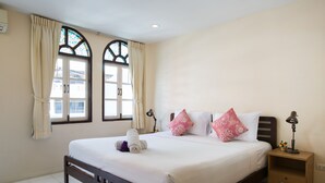 A neatly made bed with white linens, two decorative pillows, a bedside table with a lamp, and a window with curtains. - Stefans Guesthouse (Pattaya)