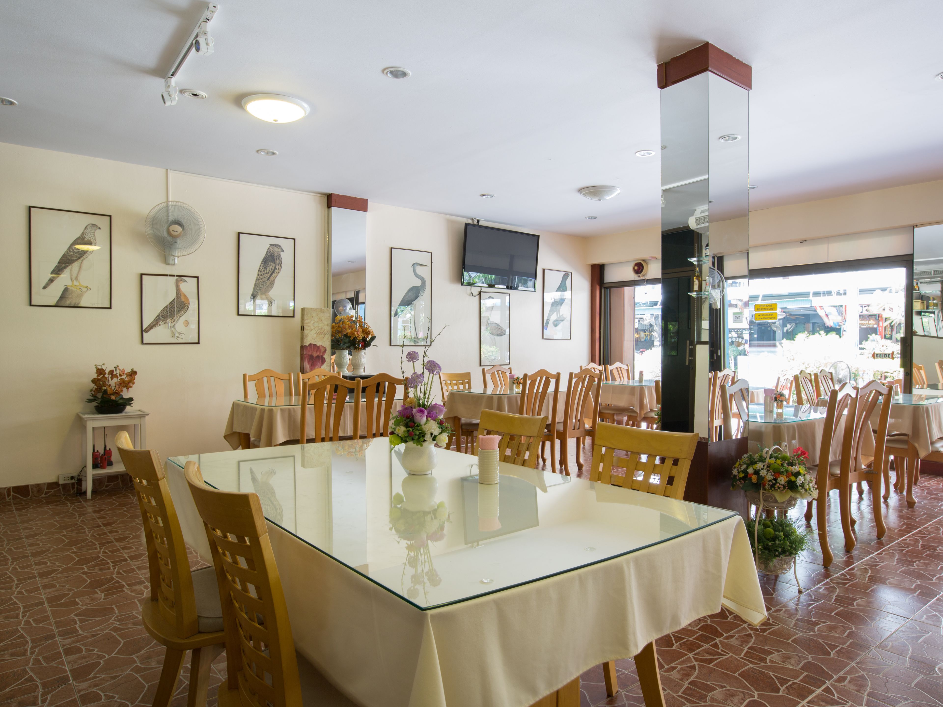 A dining area with a glass table, wooden chairs, and a white tablecloth. There are framed bird paintings on the walls and a ceiling fan.
