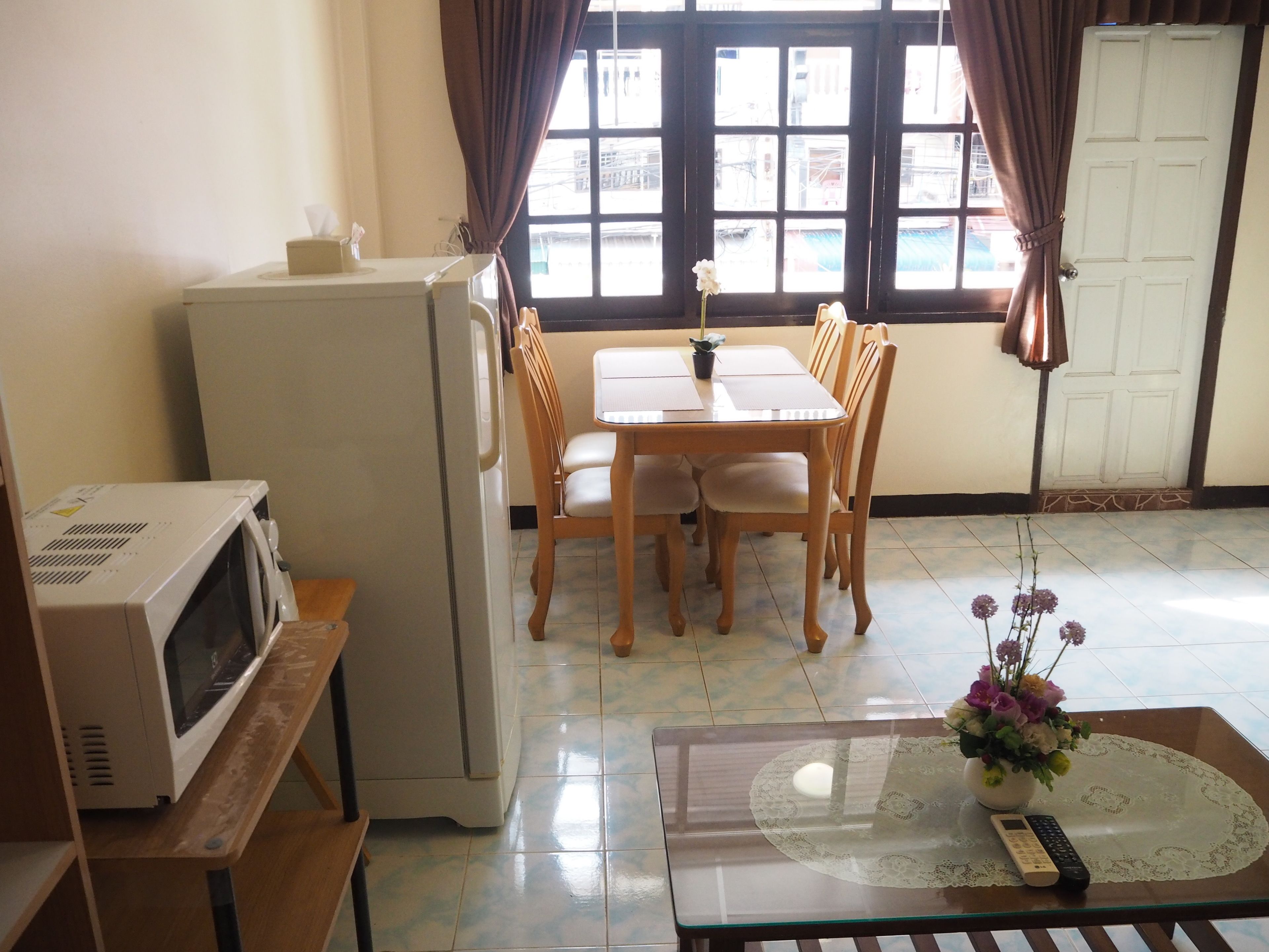 A compact living space with a kitchenette, a dining area with a table and chairs, a small refrigerator, a microwave, a glass coffee table with a remote control, and a window with curtains.