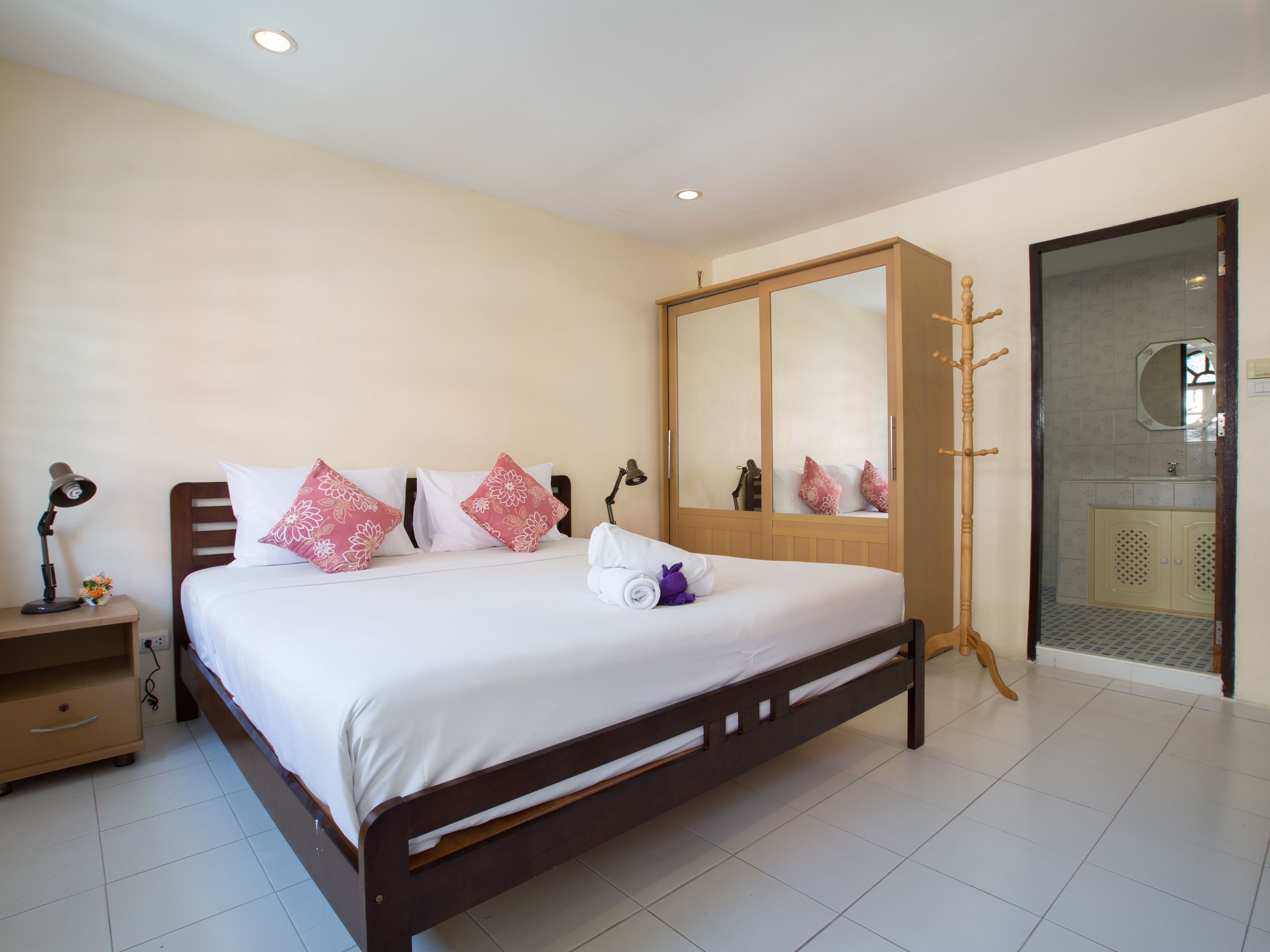 A neatly made bed with white and pink floral pillows, a wooden nightstand with a lamp, and a large wardrobe with a mirror.