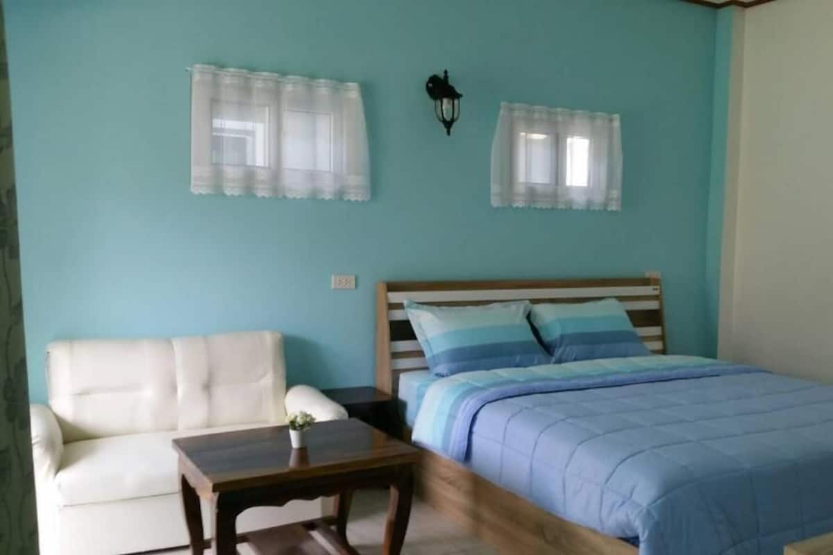 Un dormitorio con cama, mesita de noche, mesa de madera, sofá blanco y dos ventanas con cortinas traslúcidas.