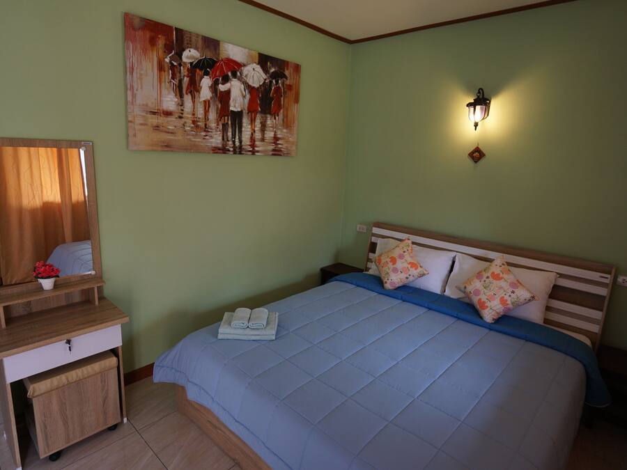 A neatly made bed with a blue quilt, a wooden nightstand with a mirror and a small plant, and a painting of people with umbrellas on the wall.