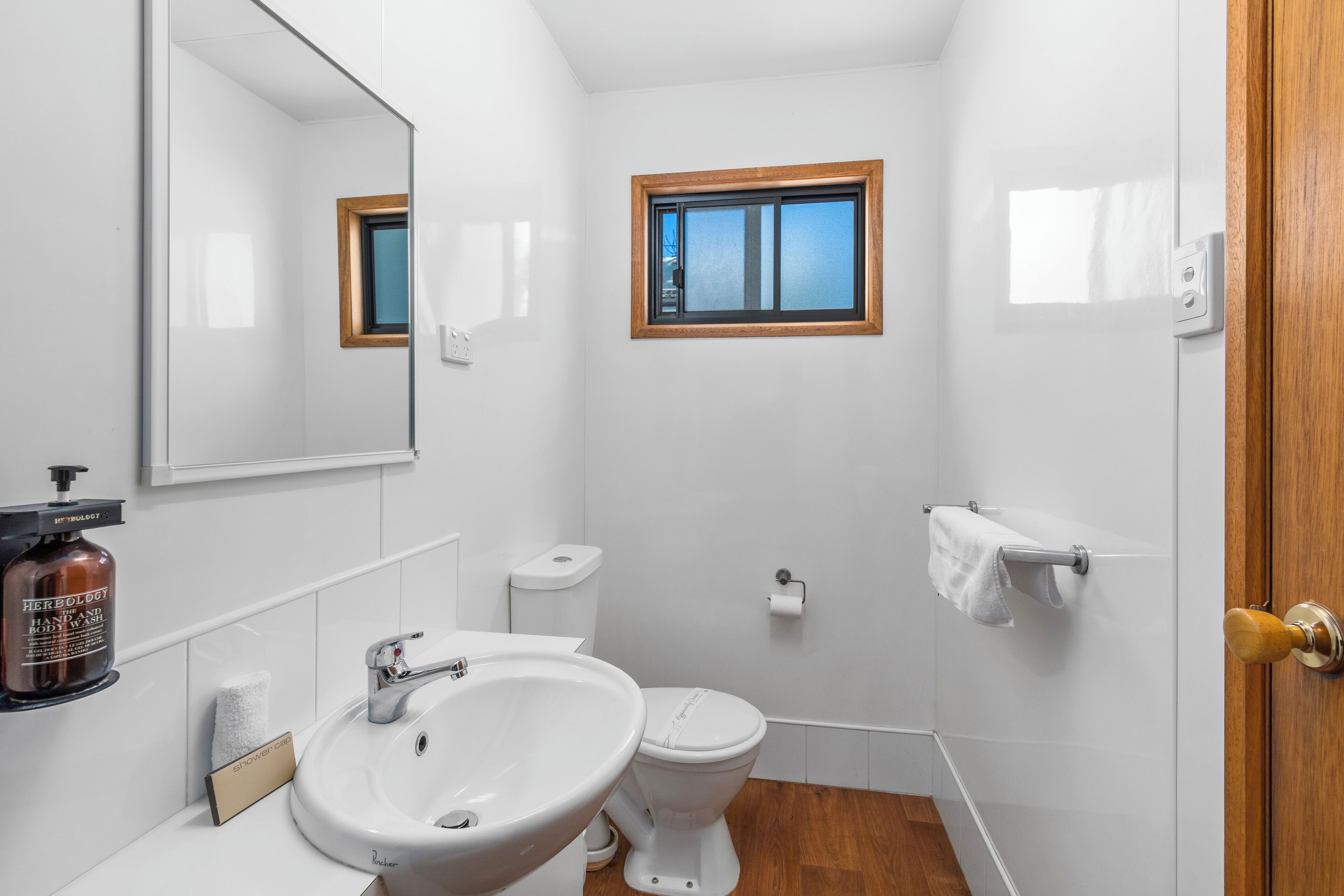Un baño con lavamanos, inodoro y espejo blancos. Se aprecia una ventana con marco de madera, un perchero y un dispensador de jabón en la pared.