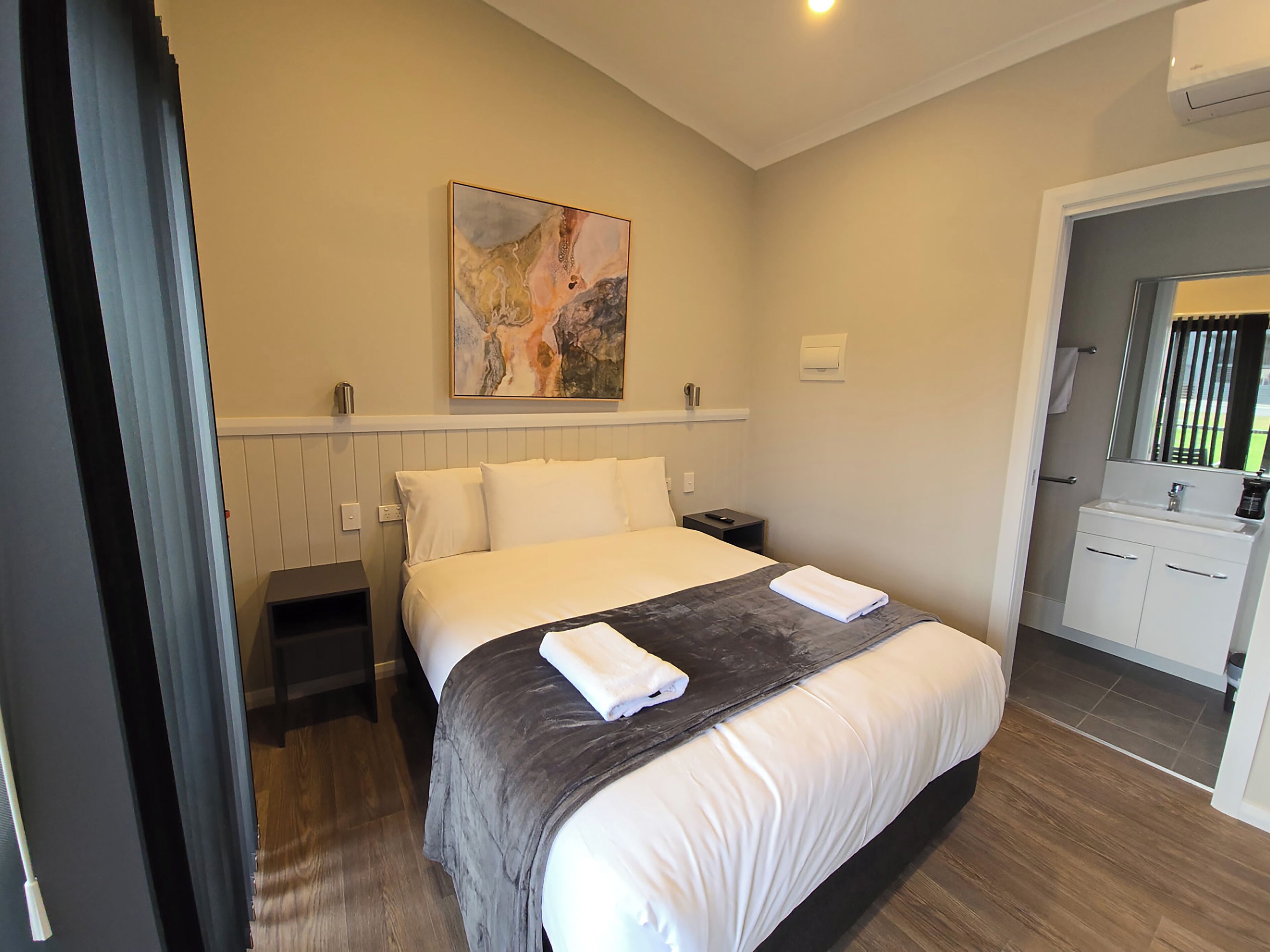 Habitació d'hotel amb un llit, una taula de nit, un quadre a la paret i un bany visible a través d'una porta oberta.