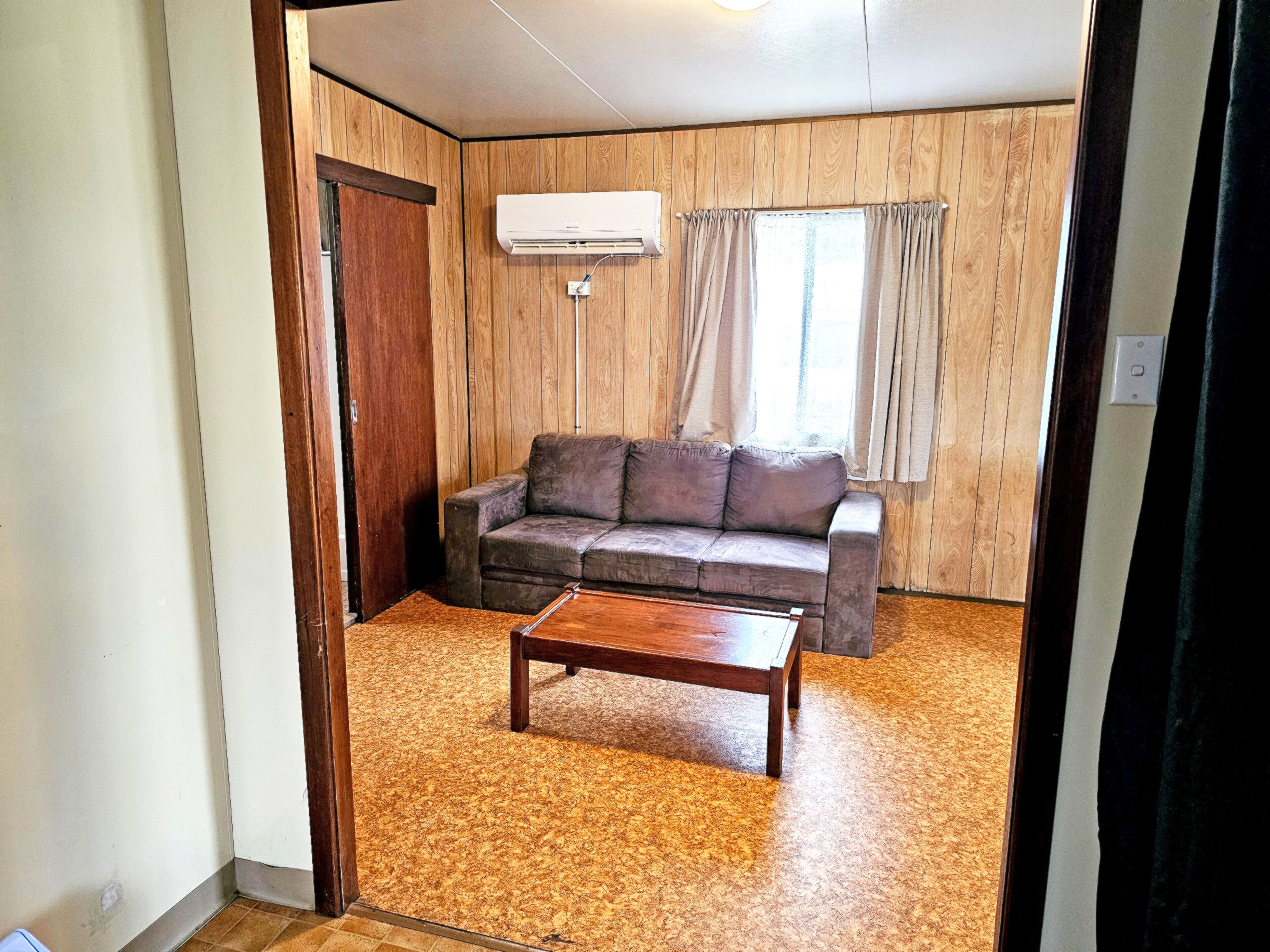 Une pièce avec un mur lambrissé en bois, un canapé gris, une table basse en bois et une fenêtre avec des rideaux.