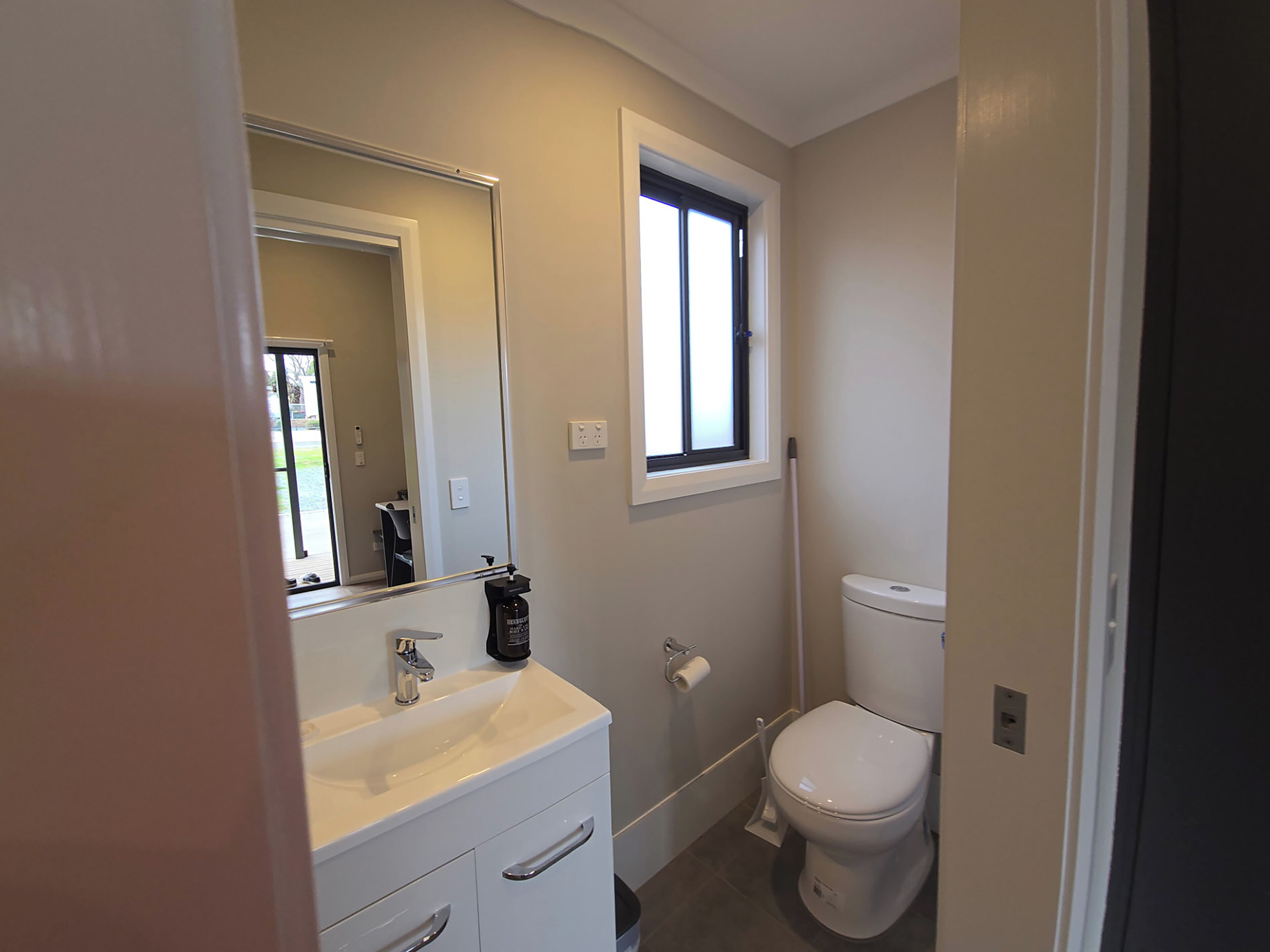 Un baño con lavamanos blanco, espejo, inodoro y ventana.