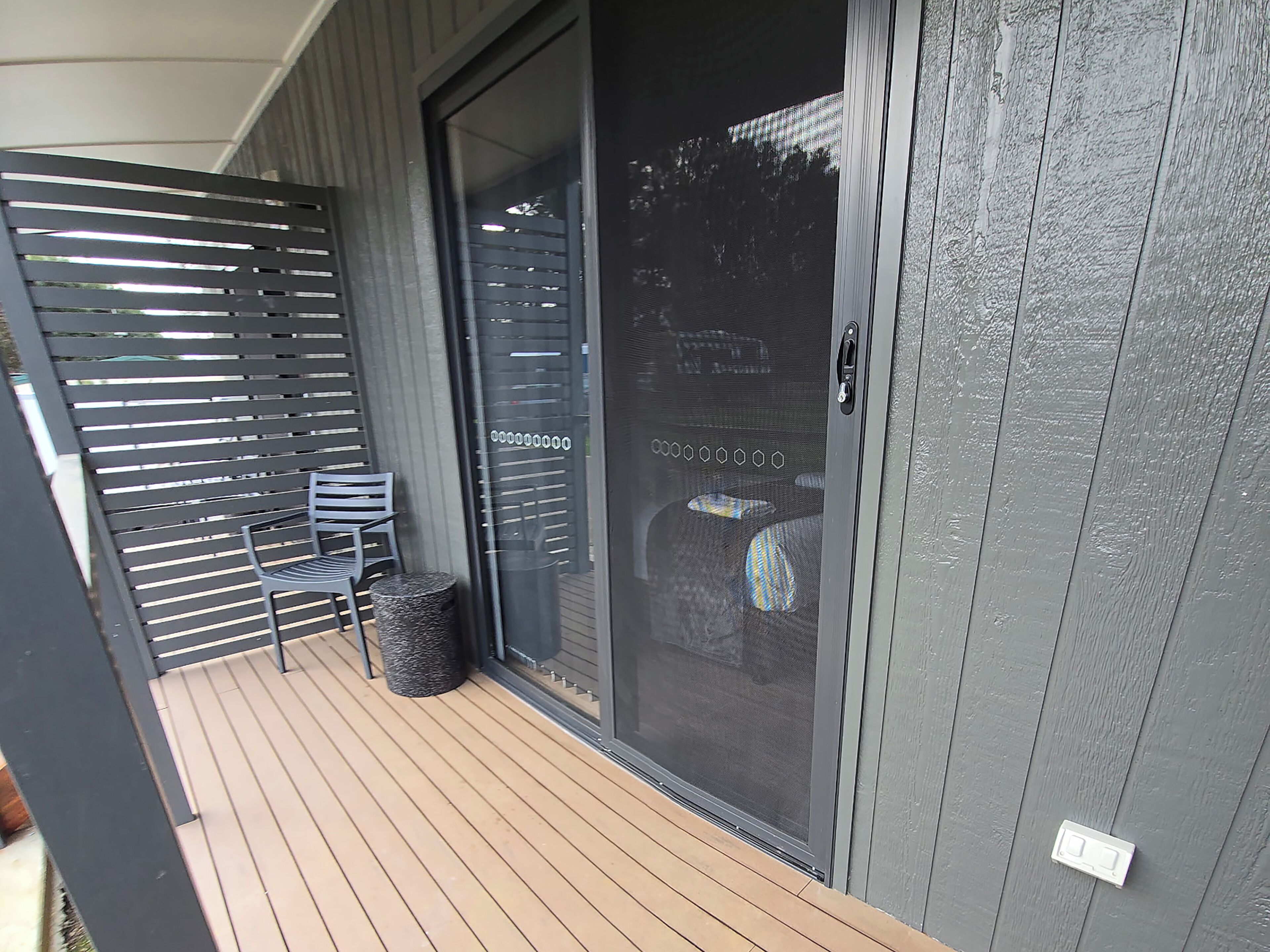 A modern building exterior with a sliding glass door, a covered outdoor seating area with chairs, and a textured wall.