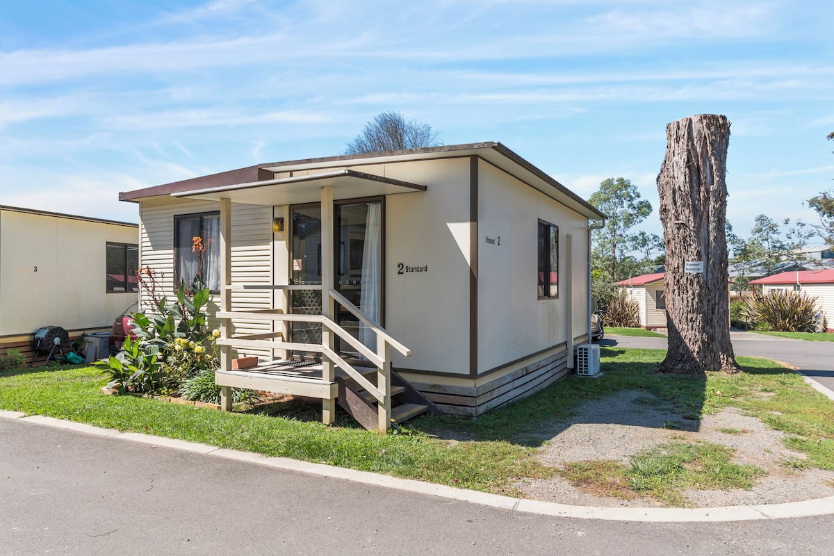 A single-story mobile home with a porch, a small garden, and a tree nearby.