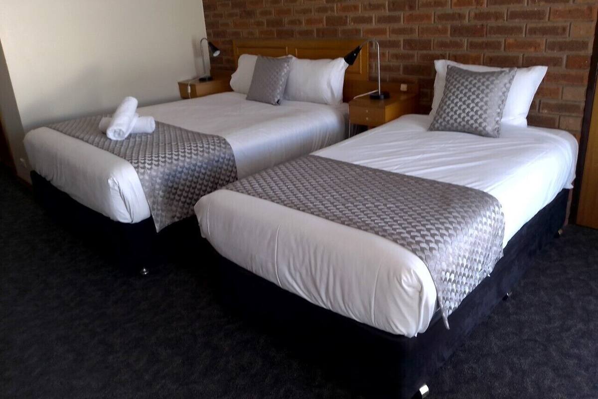 Habitación de hotel con dos camas individuales, cada una con una almohada y una toalla enrollada. La habitación tiene una pared de ladrillo y un suelo oscuro alfombrado.