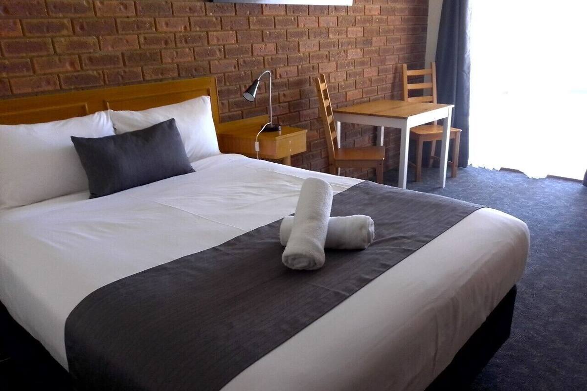 Una cama bien tendida con una colcha gris oscuro, sábanas blancas y dos toallas enrolladas. Se ve una mesita de noche de madera con una lámpara junto a la cama. En la esquina, hay una silla de madera y una pequeña mesa.