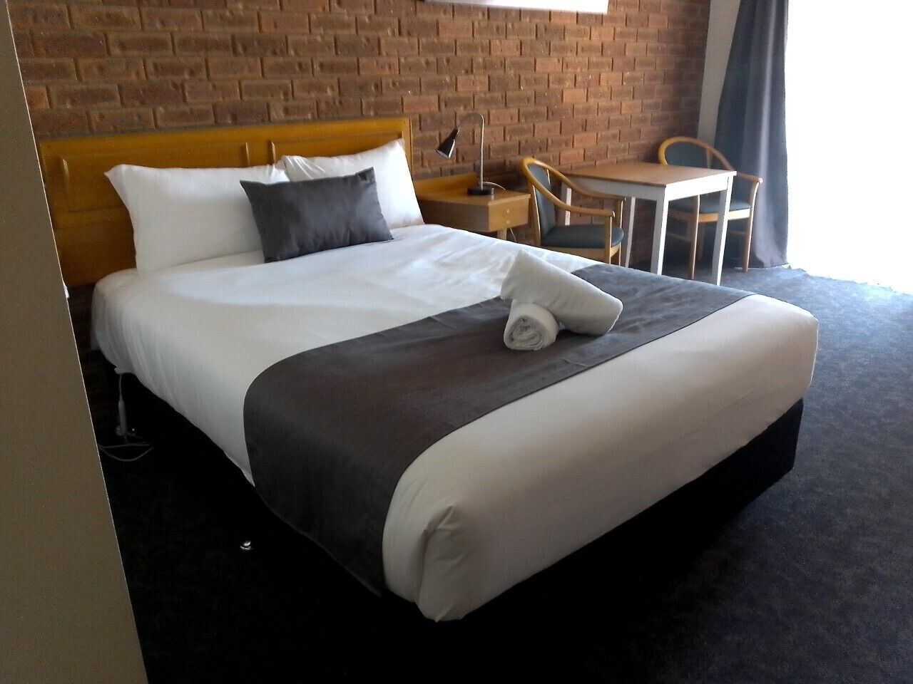 A hotel room with a large bed, a wooden headboard, a small table with a chair, and a brick wall.