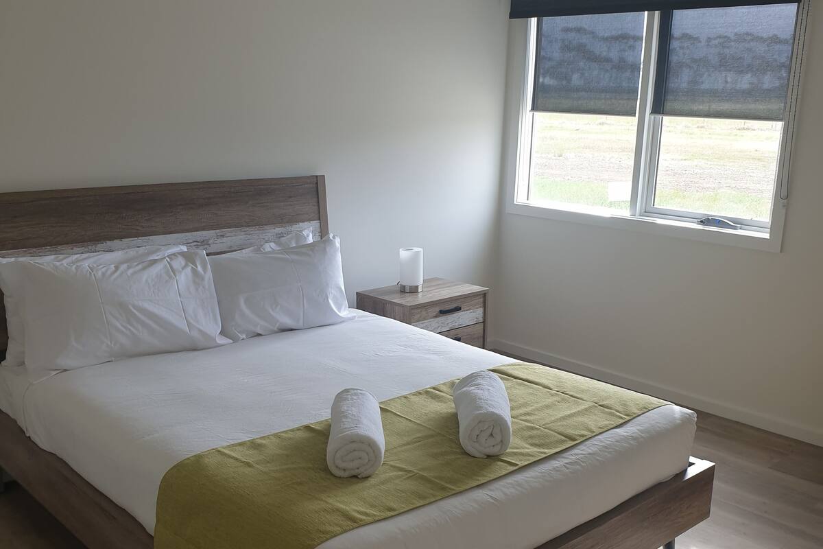 A neatly made bed with a green throw blanket and two rolled towels, a wooden headboard, a nightstand with a lamp, and a window with a view of the outdoors.