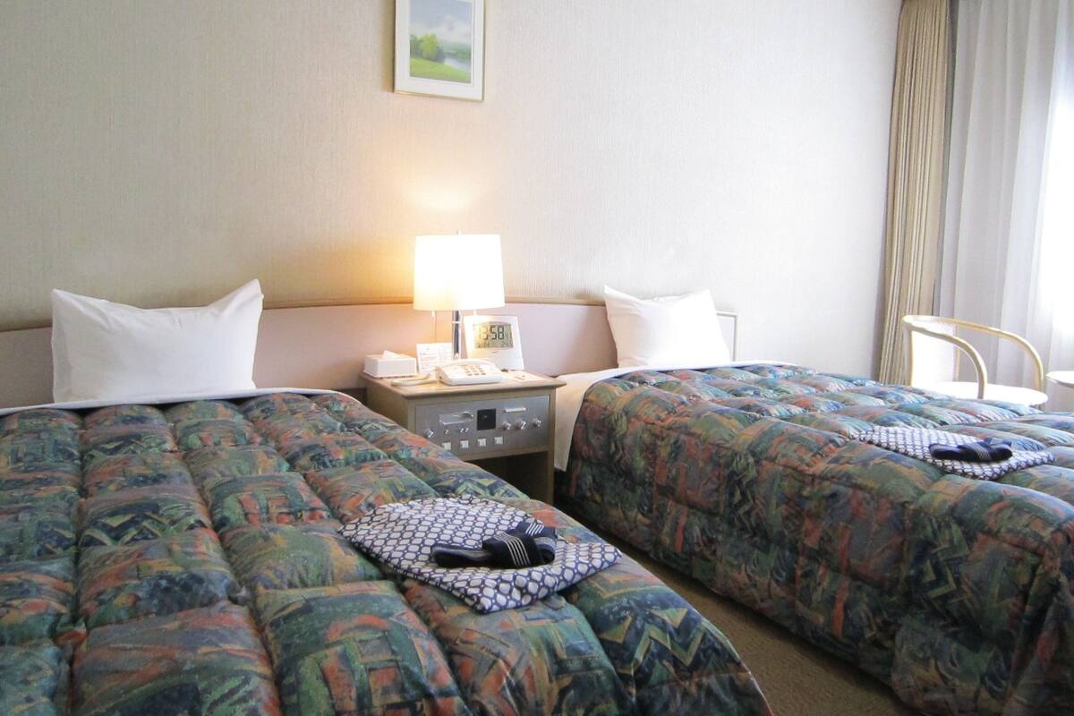 Een hotelkamer met twee bedden, een nachtkastje met een lamp, een klok en een klein tafeltje met een telefoon en zakdoekjes.