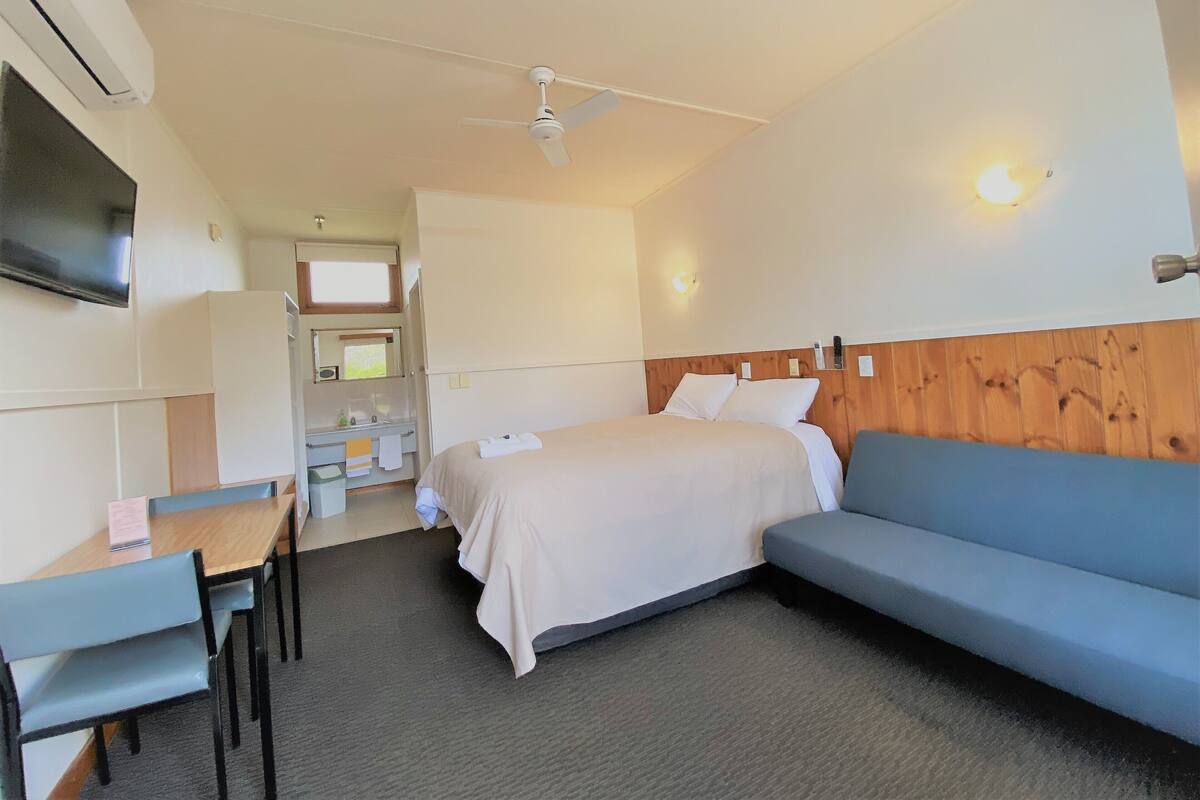 A hotel room with a bed, a sofa, a desk with chairs, a TV, a ceiling fan, and a wooden accent wall.