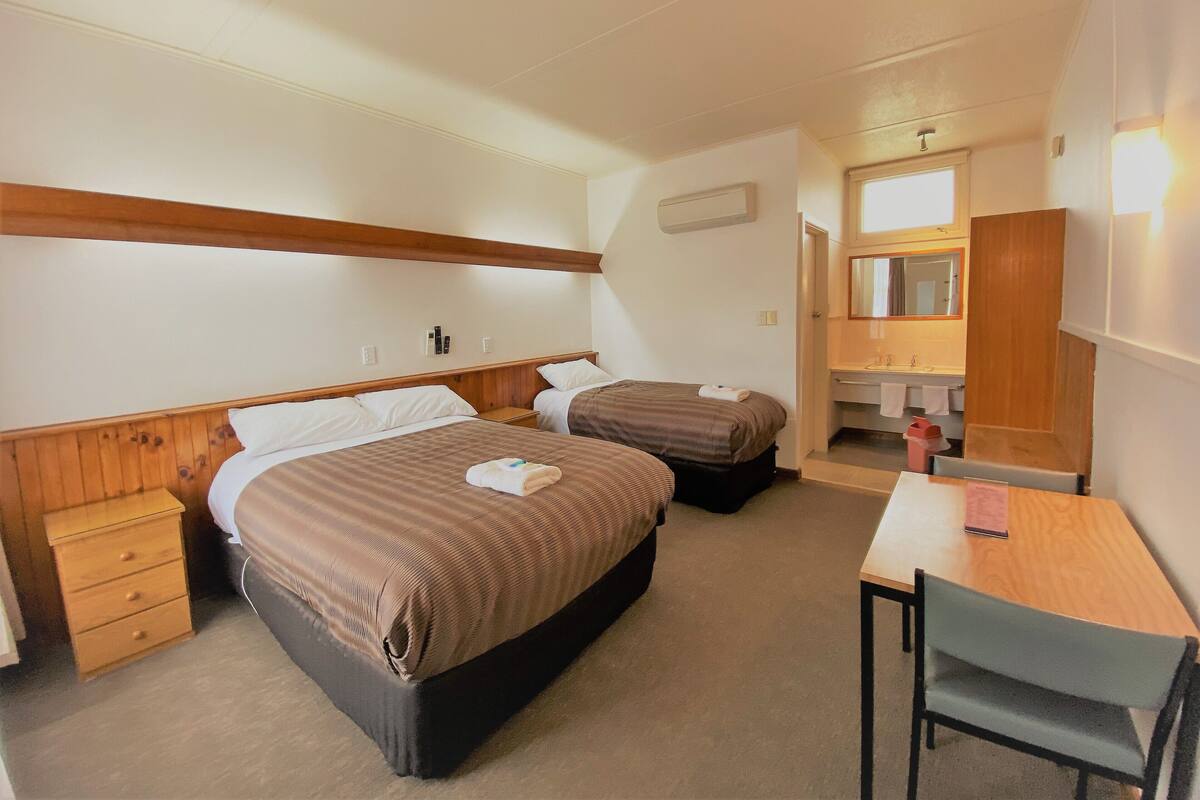 A hotel room with two beds, a desk, a chair, a small dresser, and a bathroom with a mirror and sink.