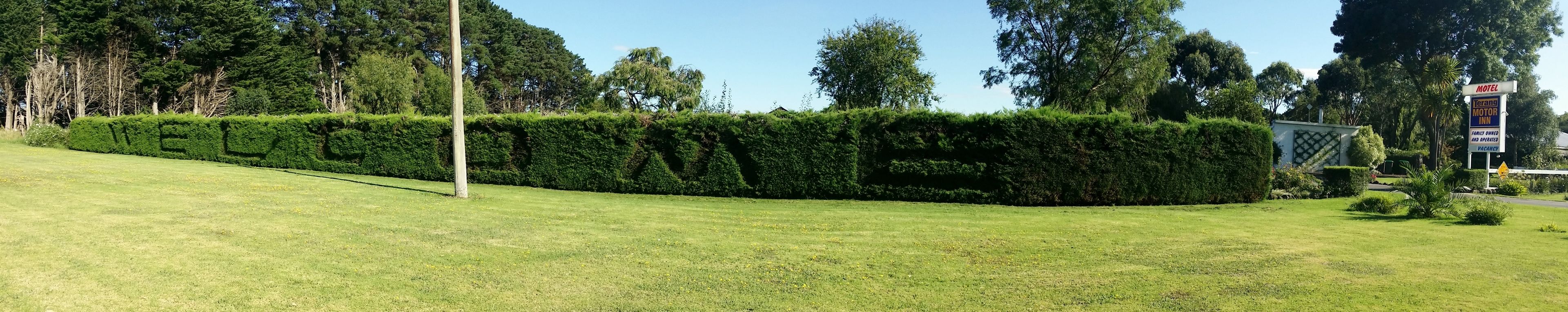 Un seto cuidado forma la palabra 'WELCOME' en un paisaje verde y exuberante, con un cartel de motel al fondo.