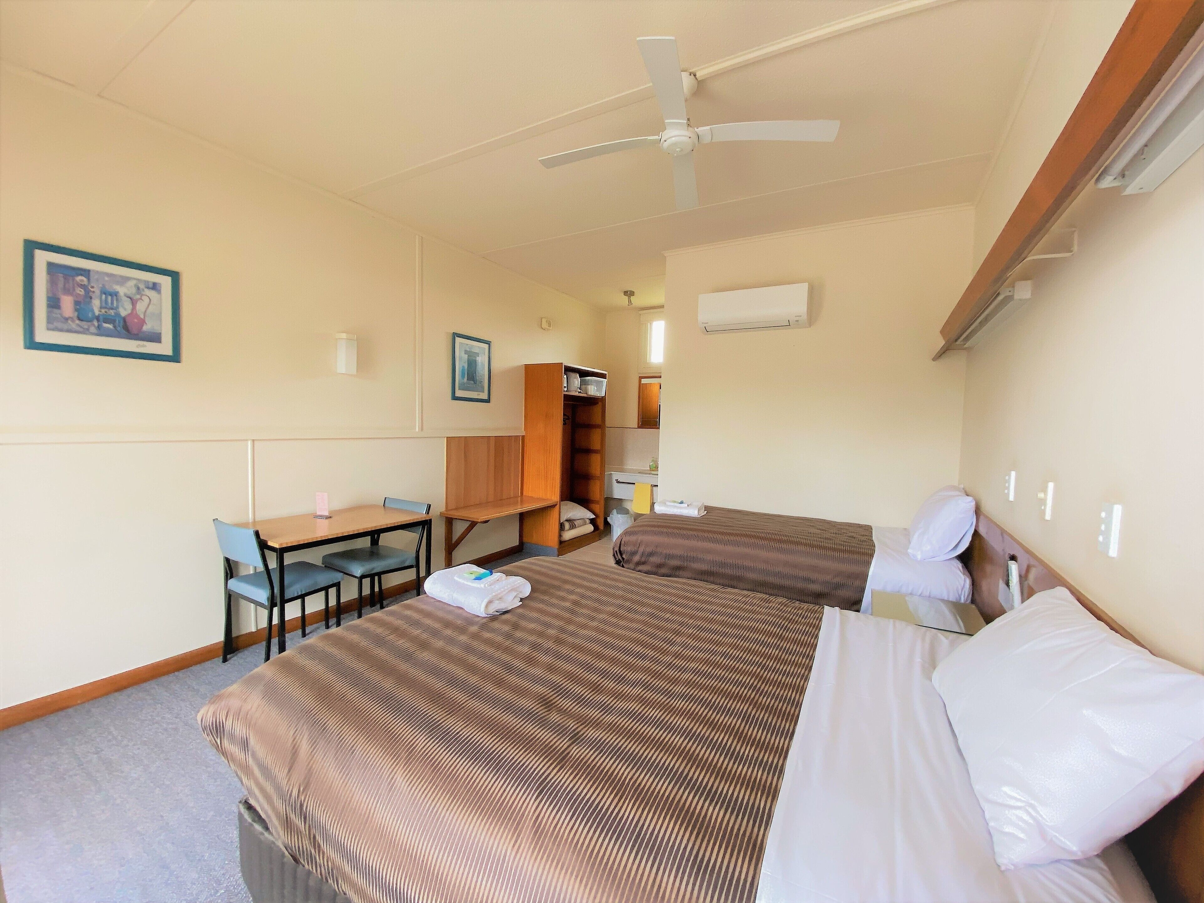 A hotel room with two beds, a desk, a chair, a wardrobe, and a ceiling fan.