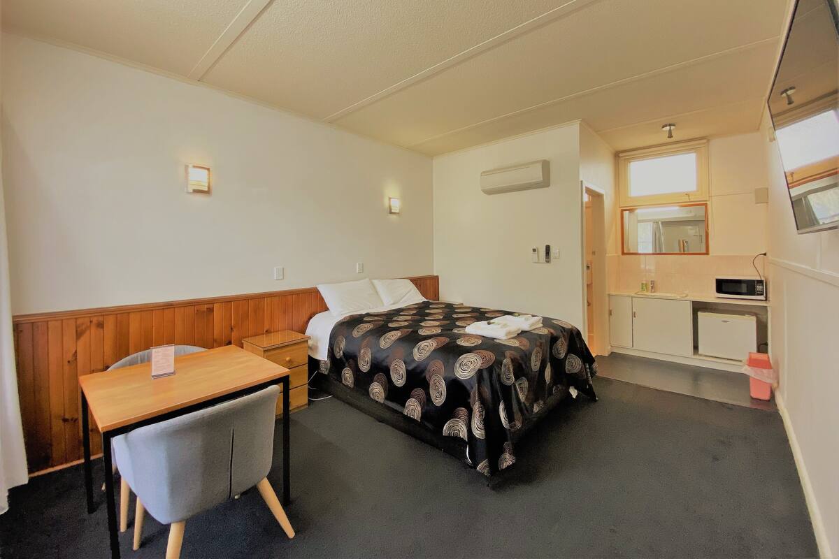 A small, clean room with a bed, a desk, a chair, a microwave, and a refrigerator.