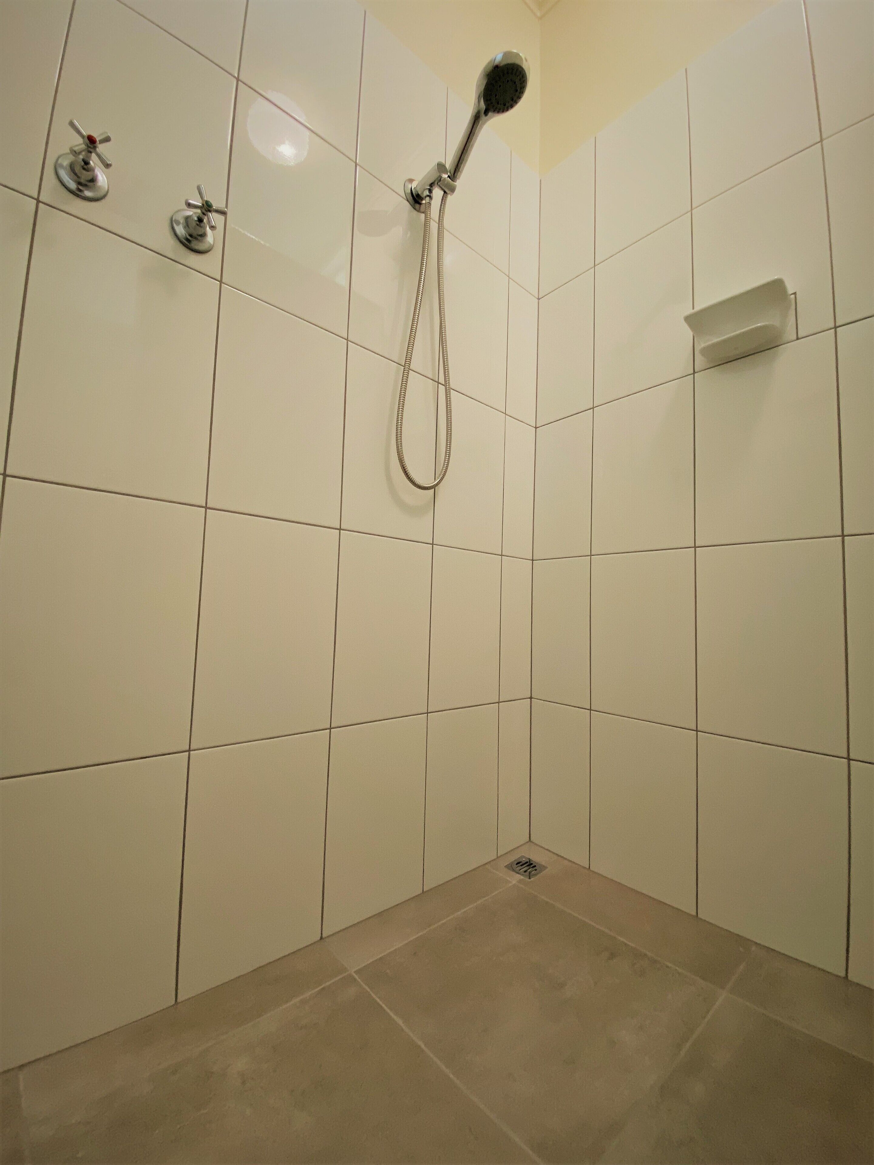 Un baño con paredes y piso de baldosas blancas, un cabezal de ducha y dos grifos.