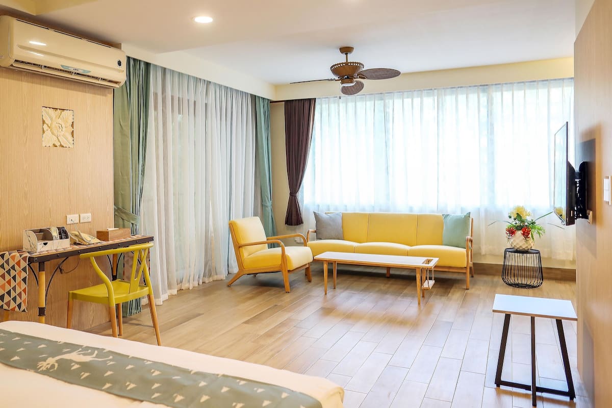 Een hotelkamer met een bed, een bureau met telefoon, een gele fauteuil, een bank, een kleine tafel en een plafondventilator.