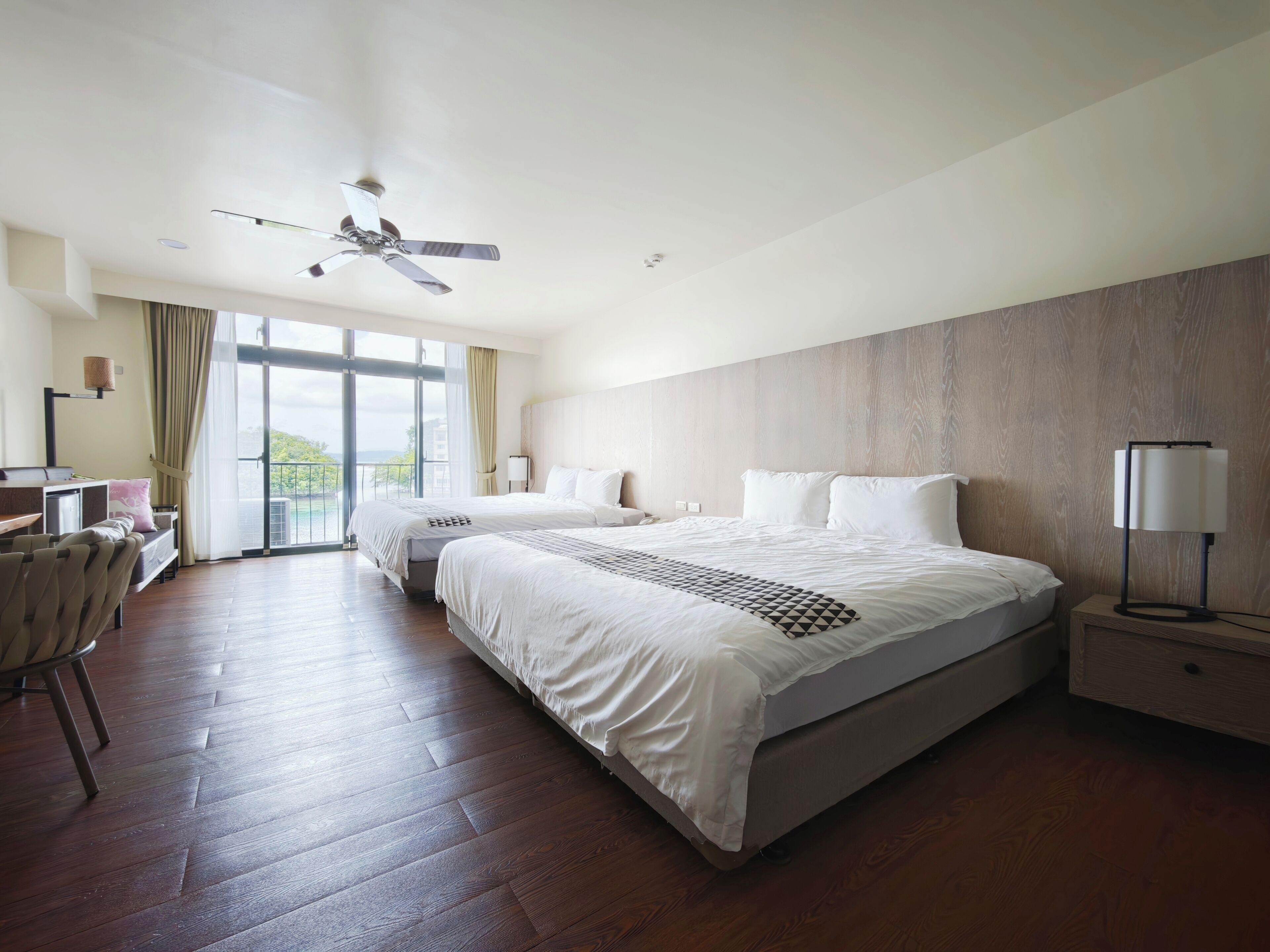 Habitación de hotel con dos camas, un escritorio, una silla, un ventilador de techo y un ventanal grande.