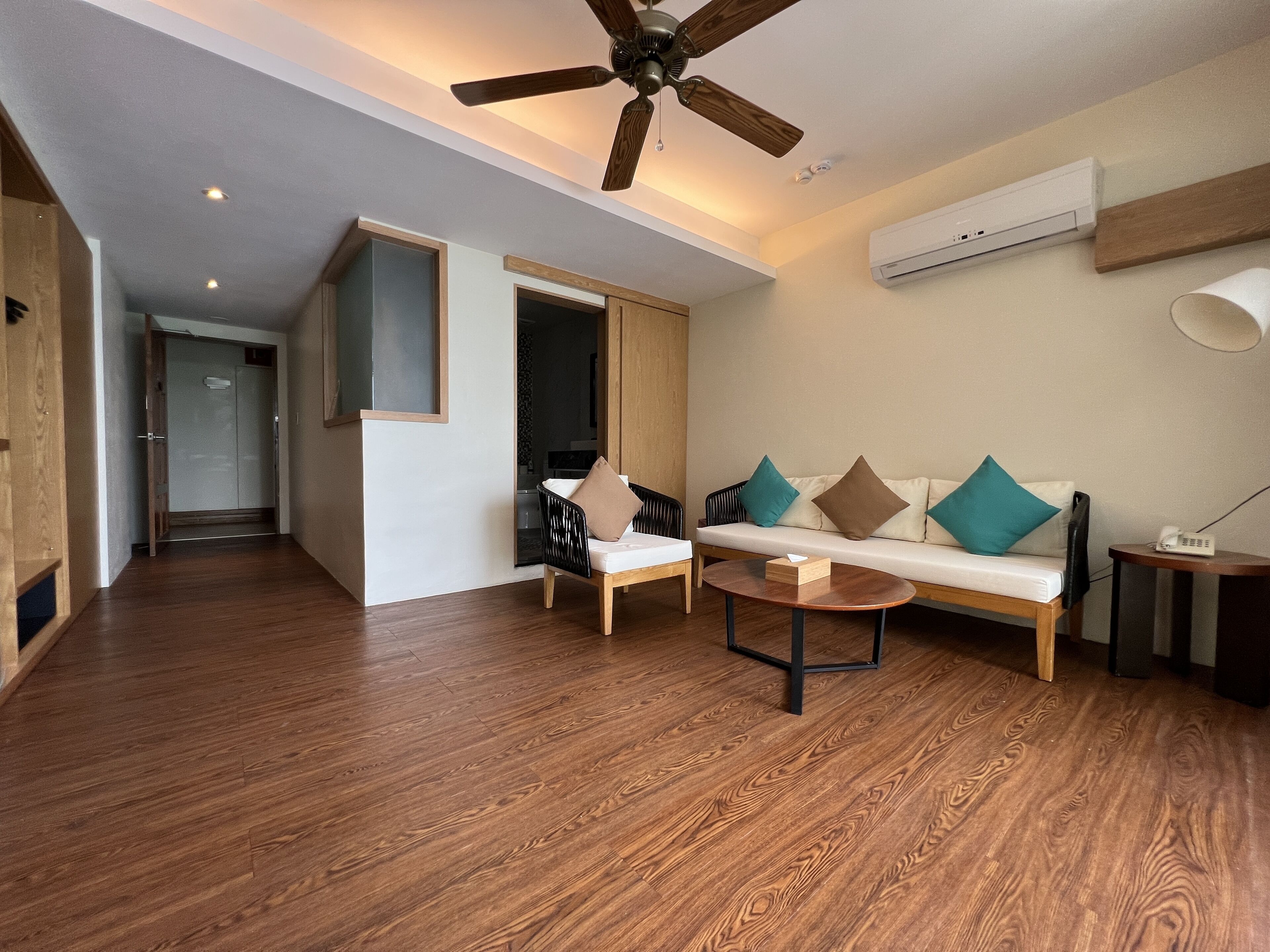 A modern living room with a wooden floor, a ceiling fan, a sofa with cushions, a coffee table, and an air conditioning unit.