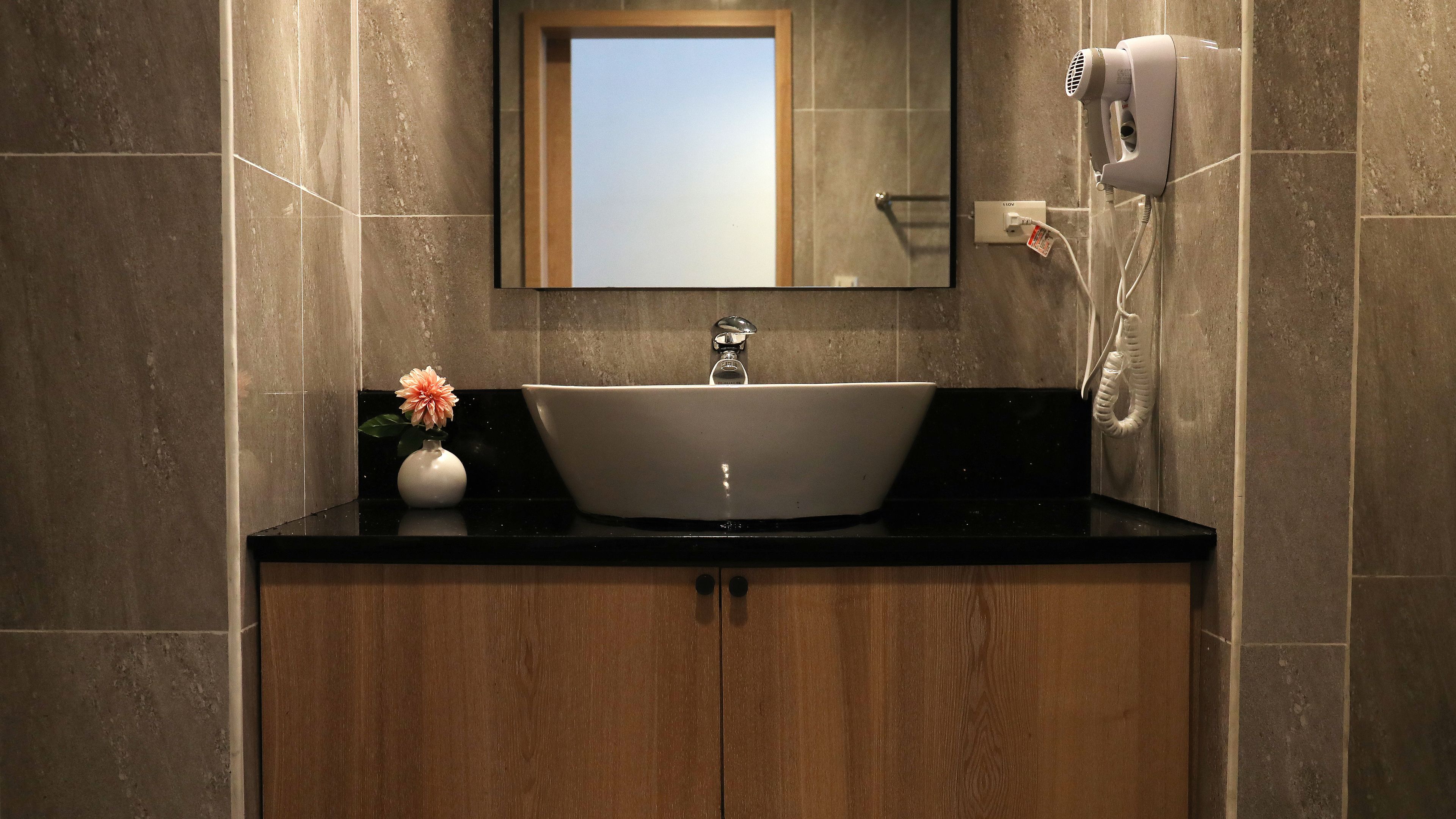 A bathroom with a sink, a mirror, a hairdryer, and a vase with a flower.