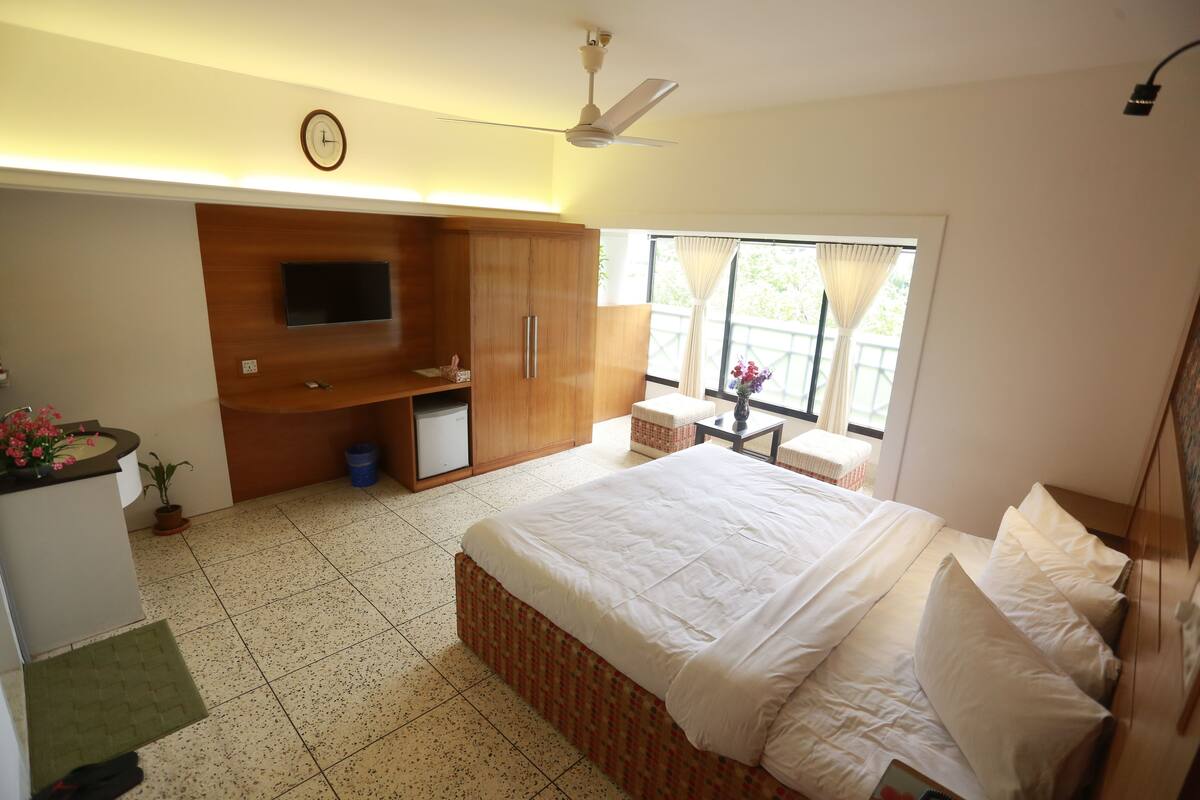 A bedroom with a bed, a TV mounted on the wall, a ceiling fan, a window with curtains, and a small table with a chair.